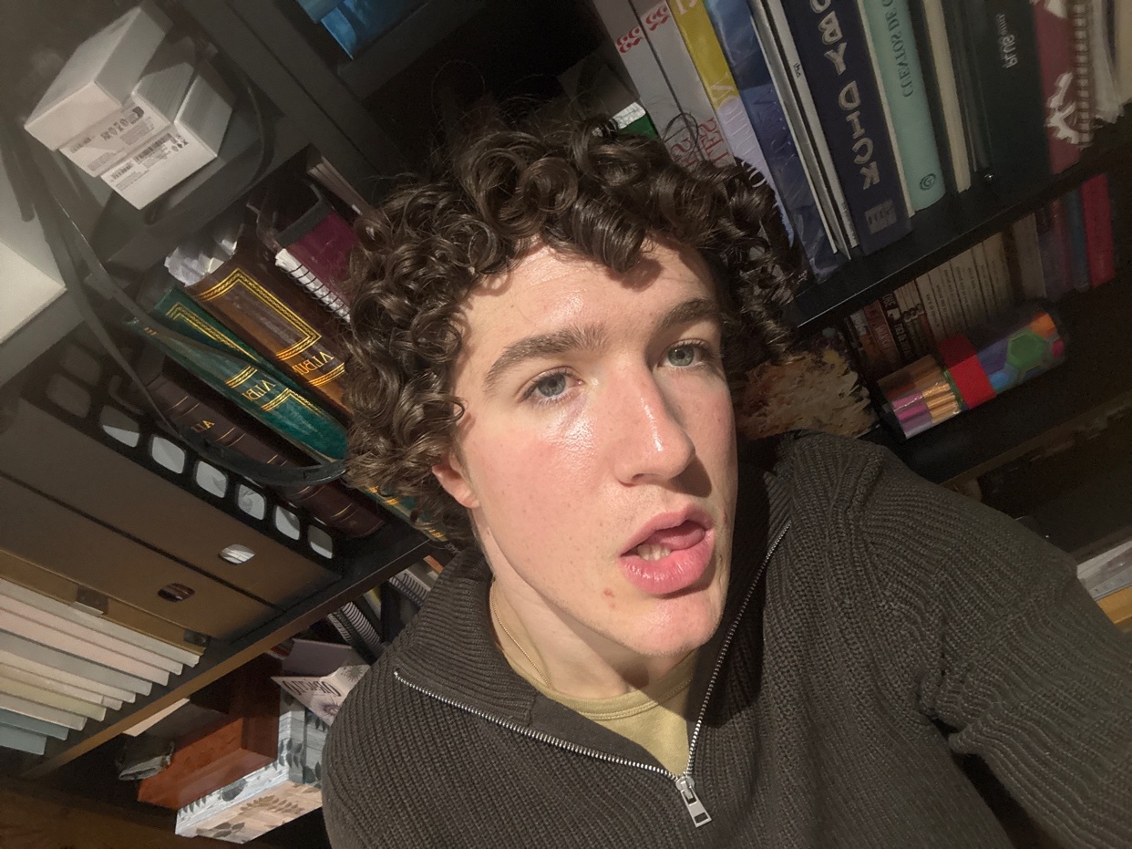 A selfie of a young man with curly hair and a slightly open mouth. He appears to be shirtless and sweaty.
