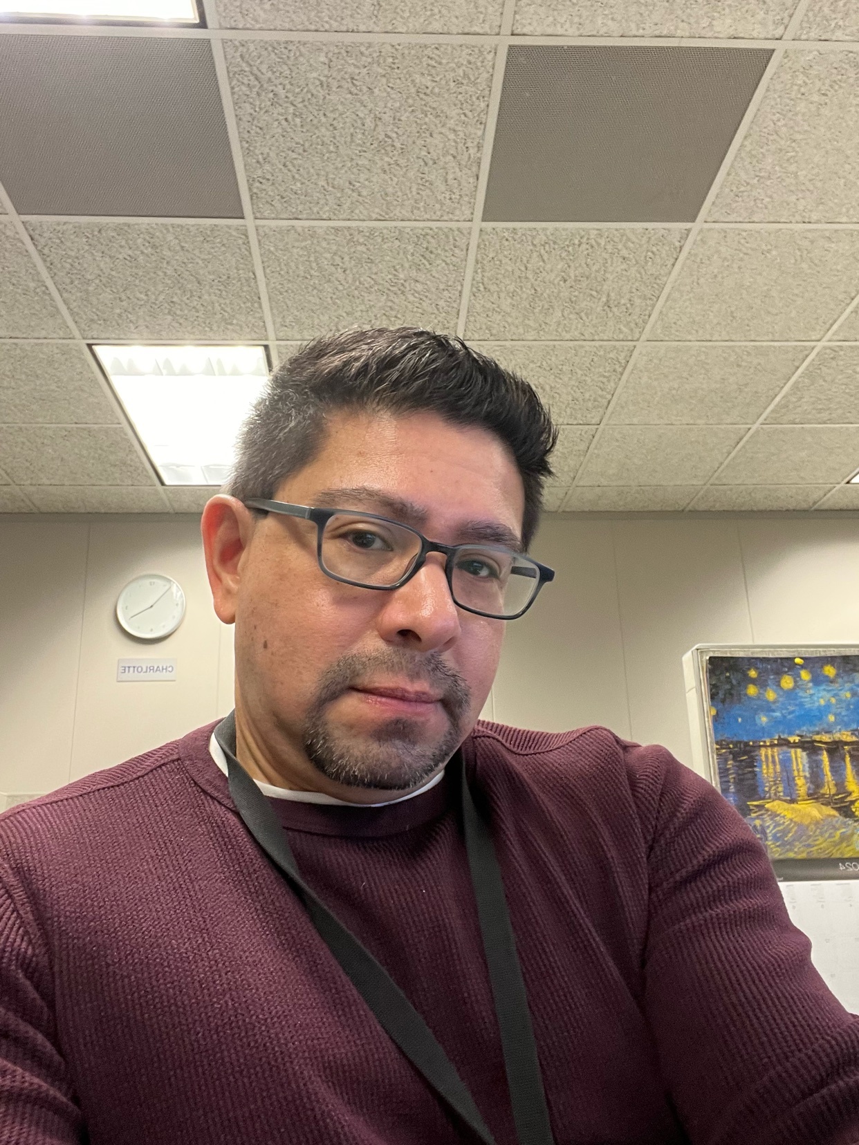 A man with glasses takes a selfie. He is wearing a maroon sweater and has short, dark hair.