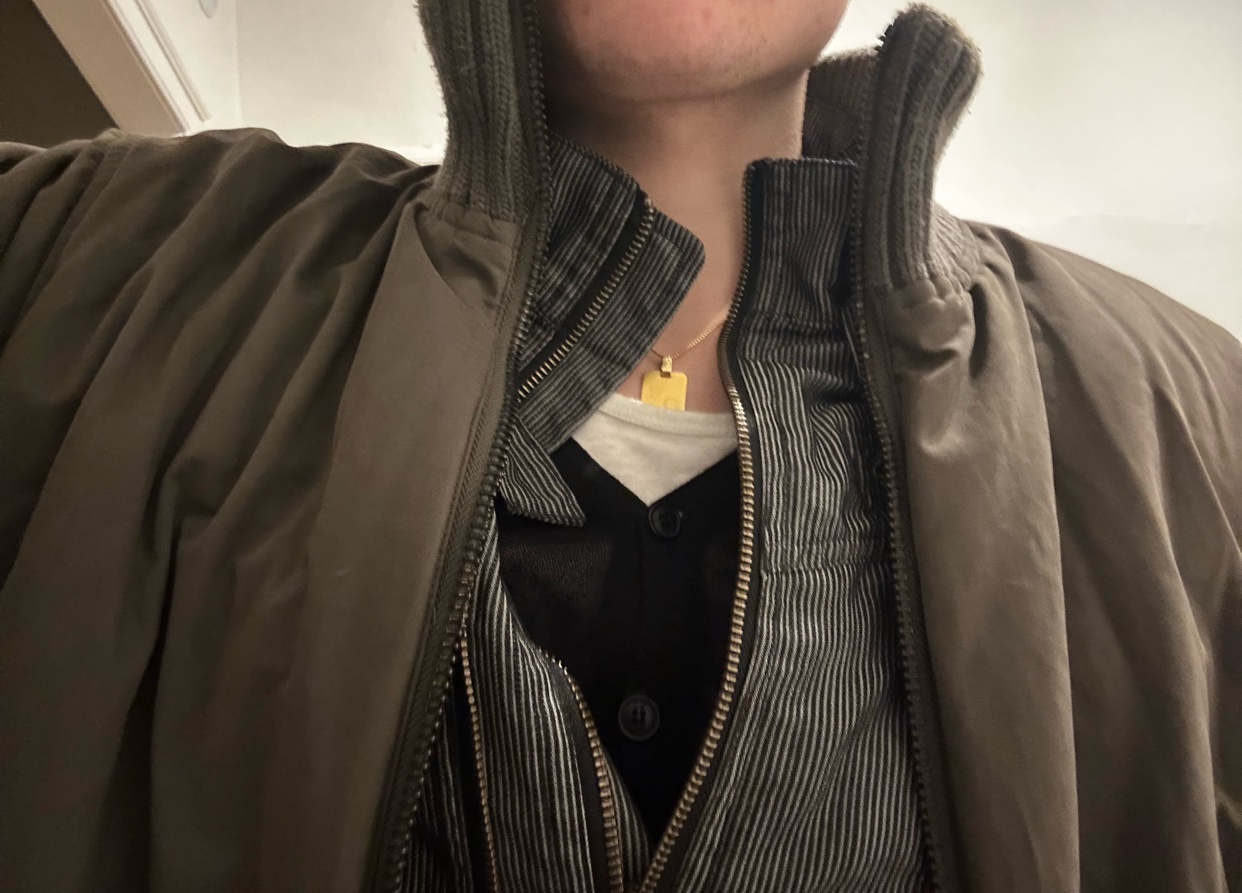 A man is wearing a stylish outfit. He is wearing a jacket, a vest, and a gold necklace.