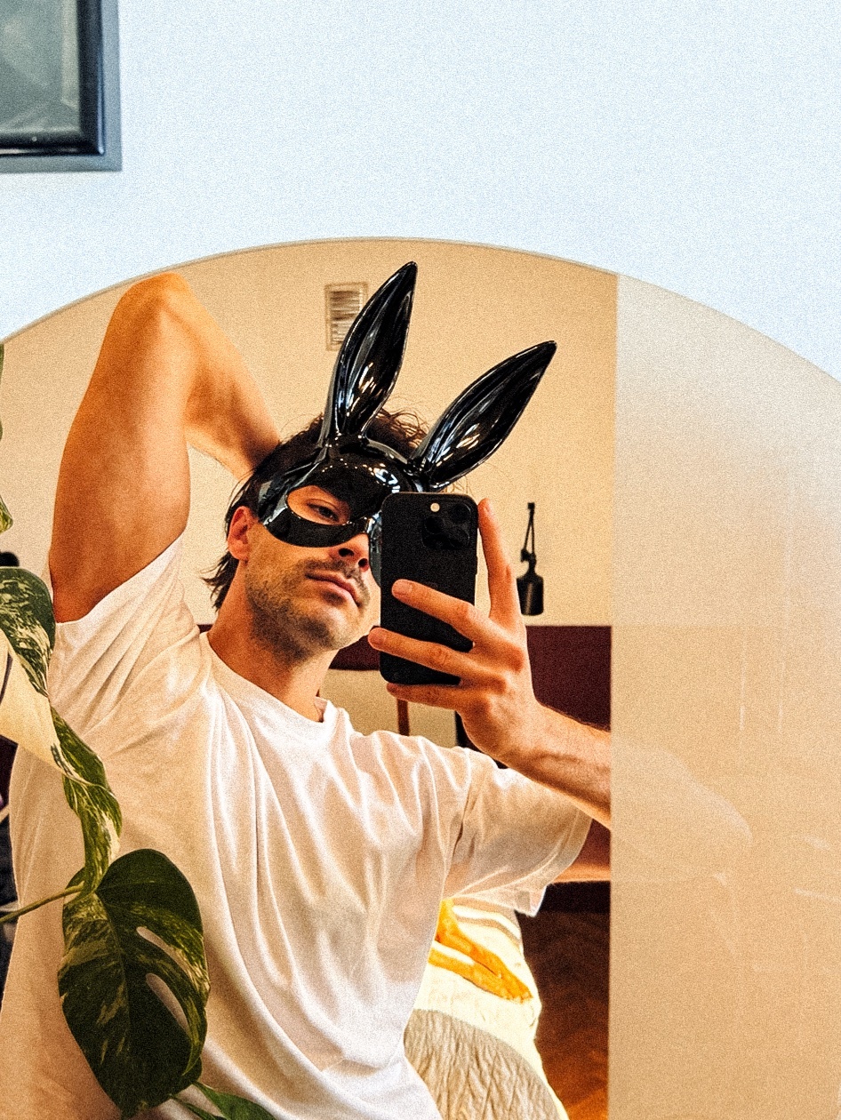 A man takes a mirror selfie while wearing a black bunny mask and a white t-shirt. He poses playfully with his arm behind his head.