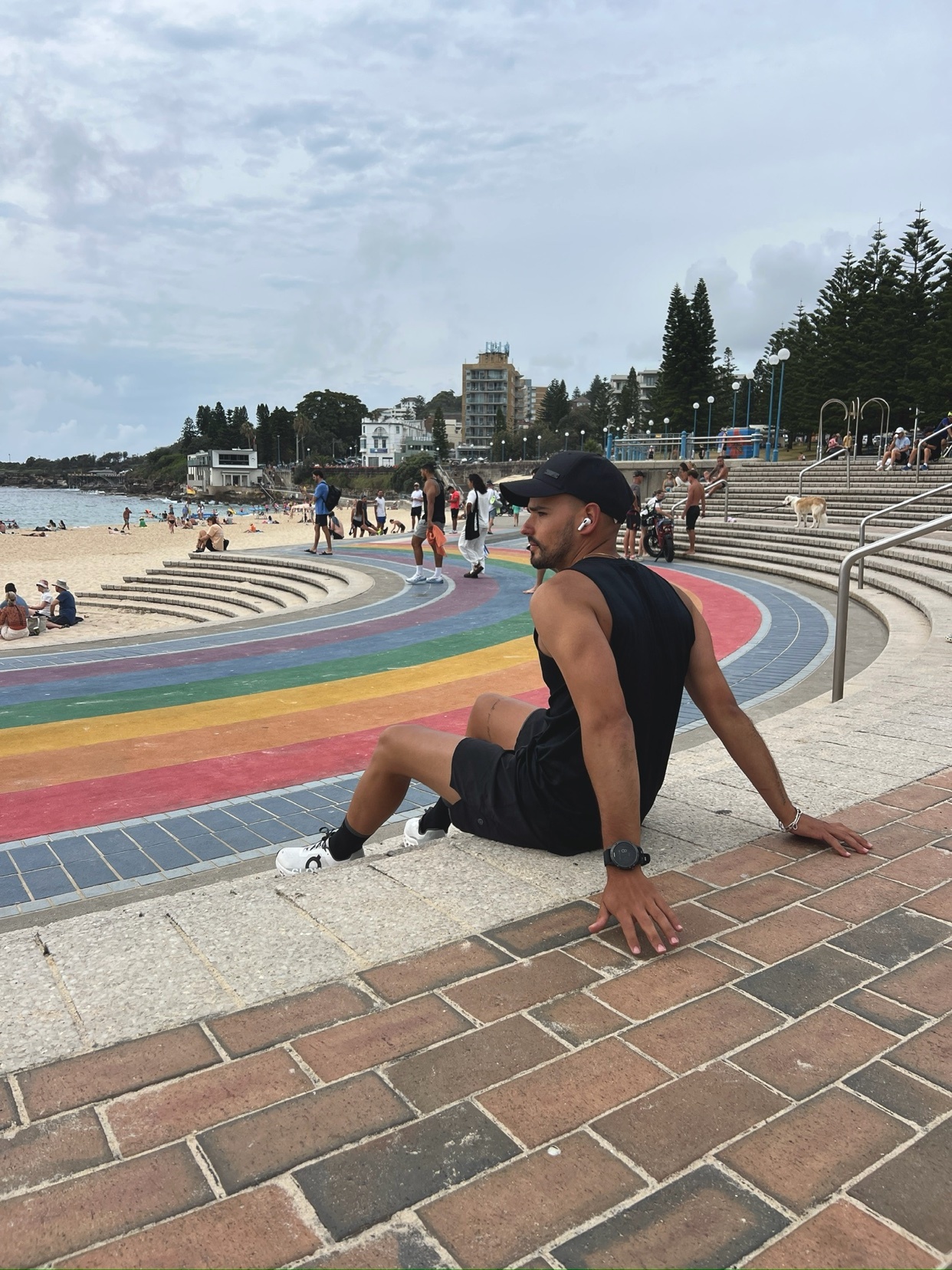 A man sits on a colorful walkway near a beach. He is shirtless and wearing athletic shorts and a cap.