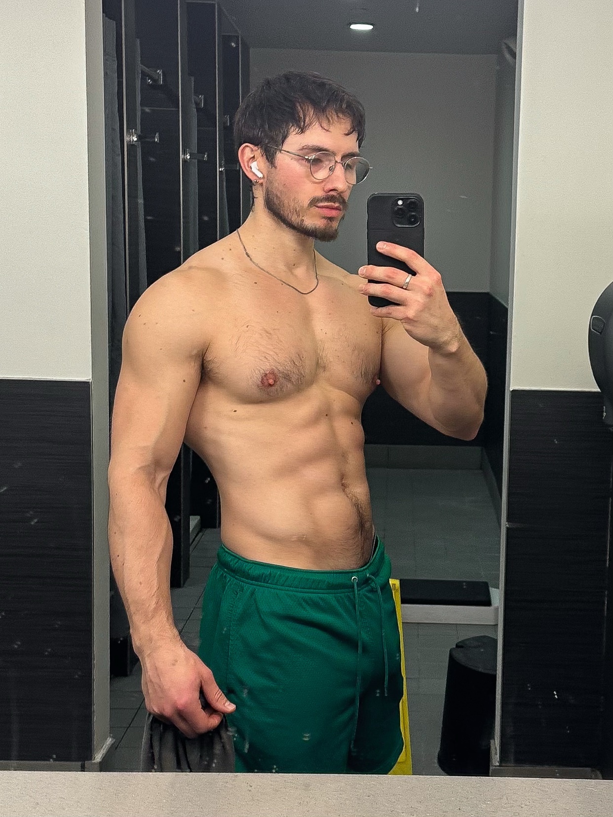A man takes a mirror selfie in a gym, showcasing his muscular physique. He is shirtless, wearing green shorts and glasses.