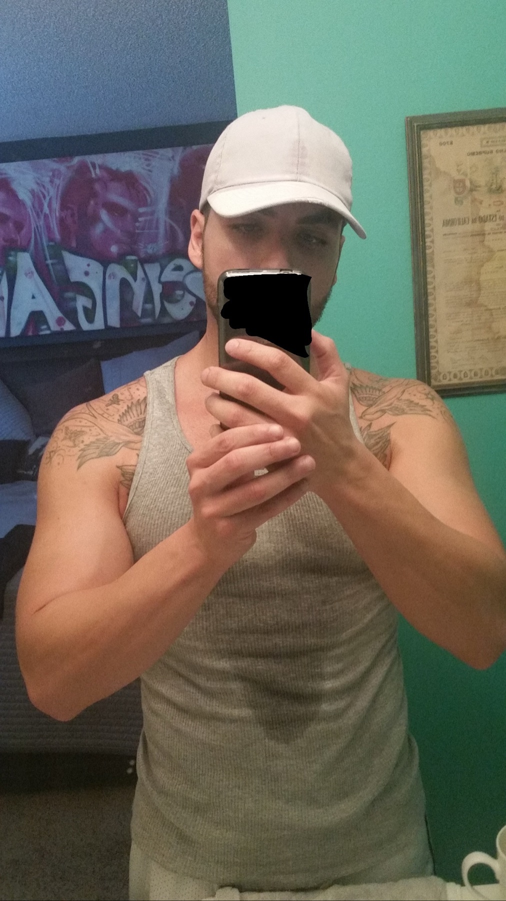 A man in a grey tank top and a white baseball cap takes a selfie in a bathroom. He has tattoos on his arms and appears to be muscular.
