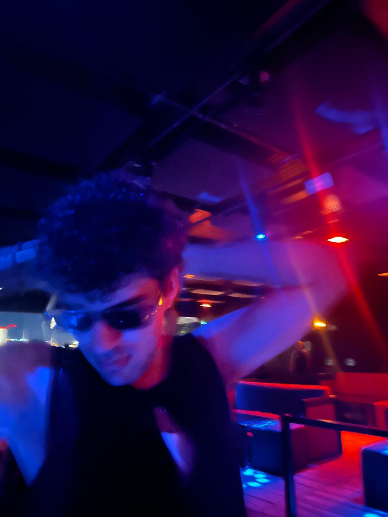 A man wearing sunglasses and a dark tank top dances in a nightclub with blue and red lights illuminating the scene.