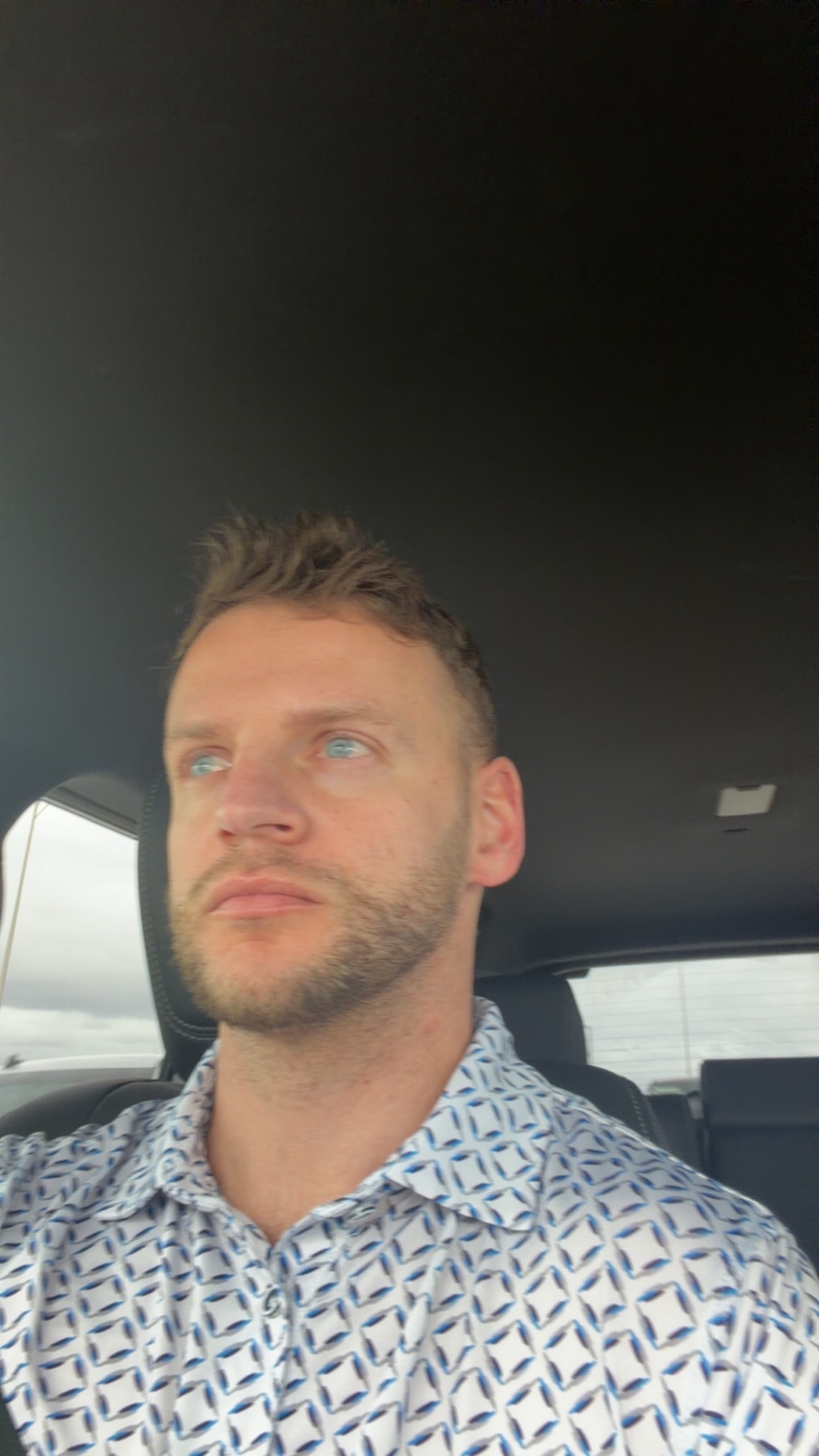 A man with short brown hair and stubble is looking up and to the side while wearing a patterned button-up shirt. The photo appears to be a selfie taken inside a vehicle.