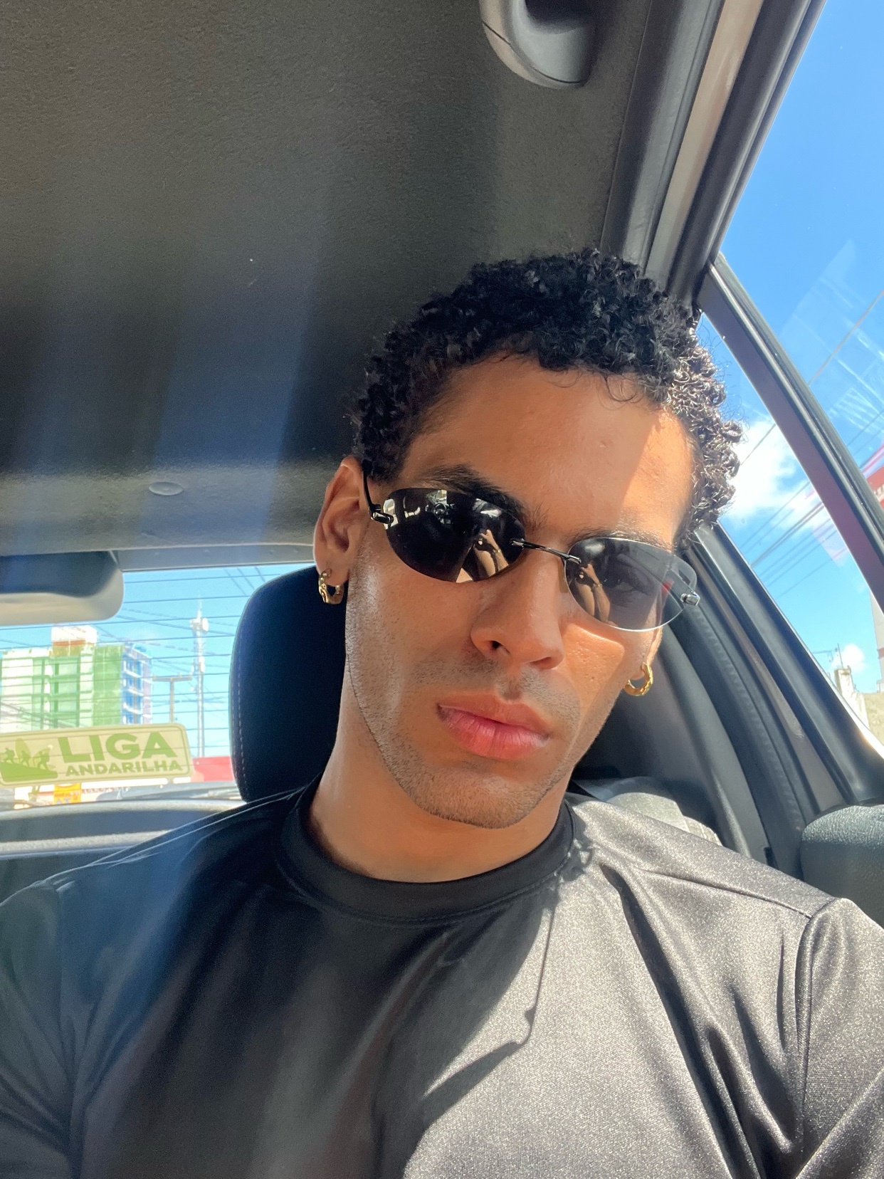 A man with curly hair and sunglasses takes a selfie in a car.