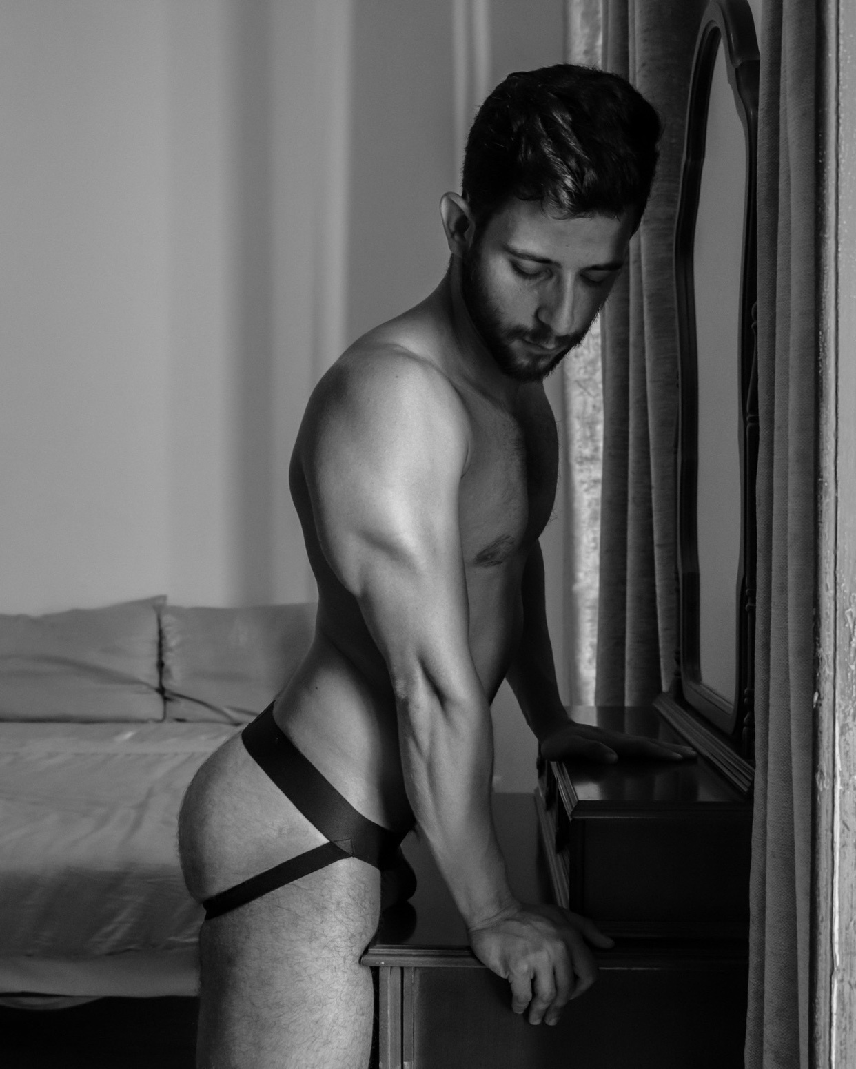 A black and white photo of a man leaning against a dresser, shirtless and wearing a jockstrap. The image has a modern aesthetic.