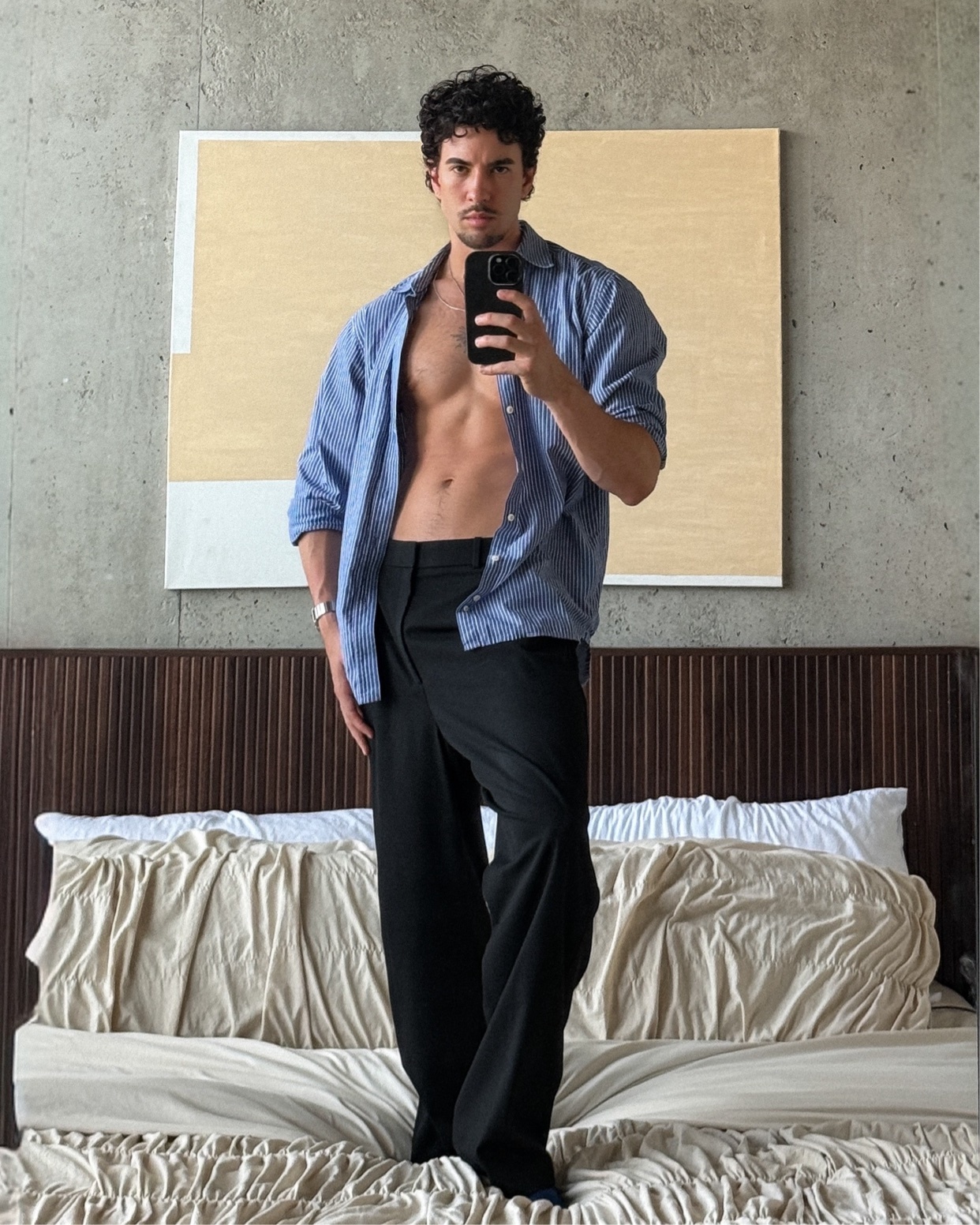 A man with curly hair poses for a selfie in a bedroom, wearing an unbuttoned shirt and black pants.