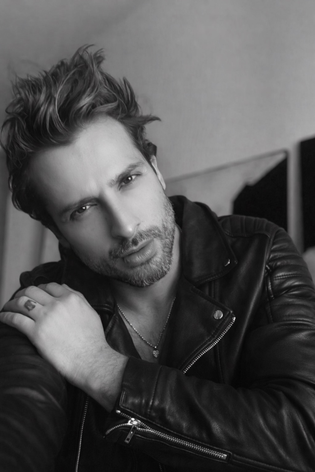 A black and white portrait of a man with stubble wearing a leather jacket and a necklace. He has a tattoo on his left hand.