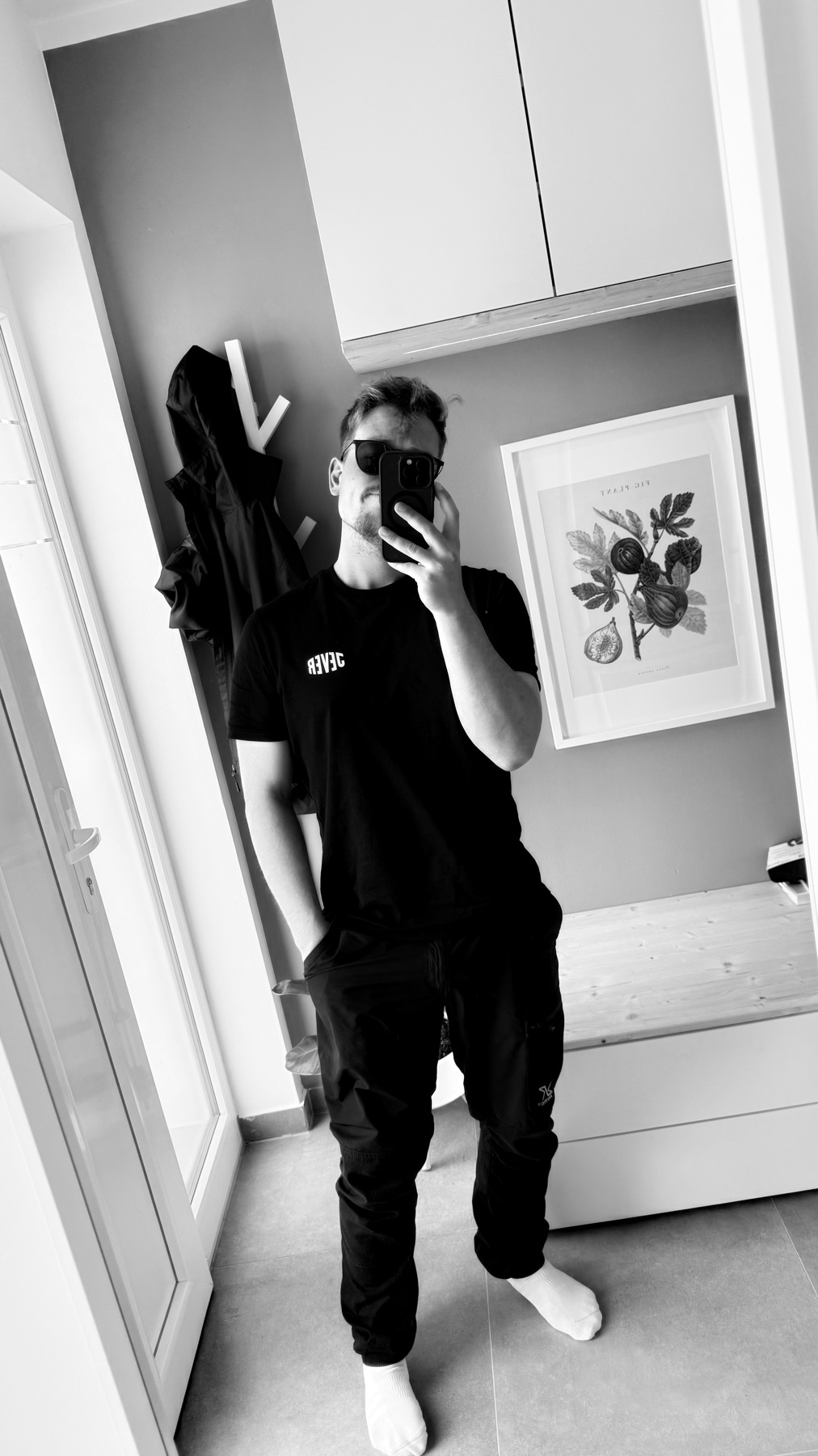 A man takes a selfie in a black t-shirt and pants, wearing sunglasses. The black and white photo shows him standing in a hallway with a framed picture on the wall.
