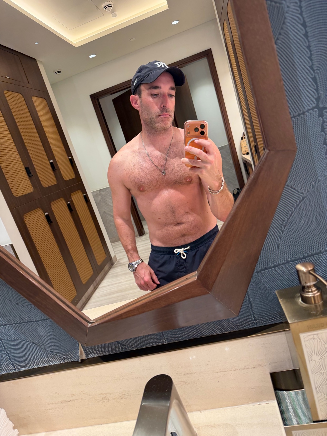 A man takes a mirror selfie, shirtless, in a bathroom. He is wearing a cap and swim trunks.