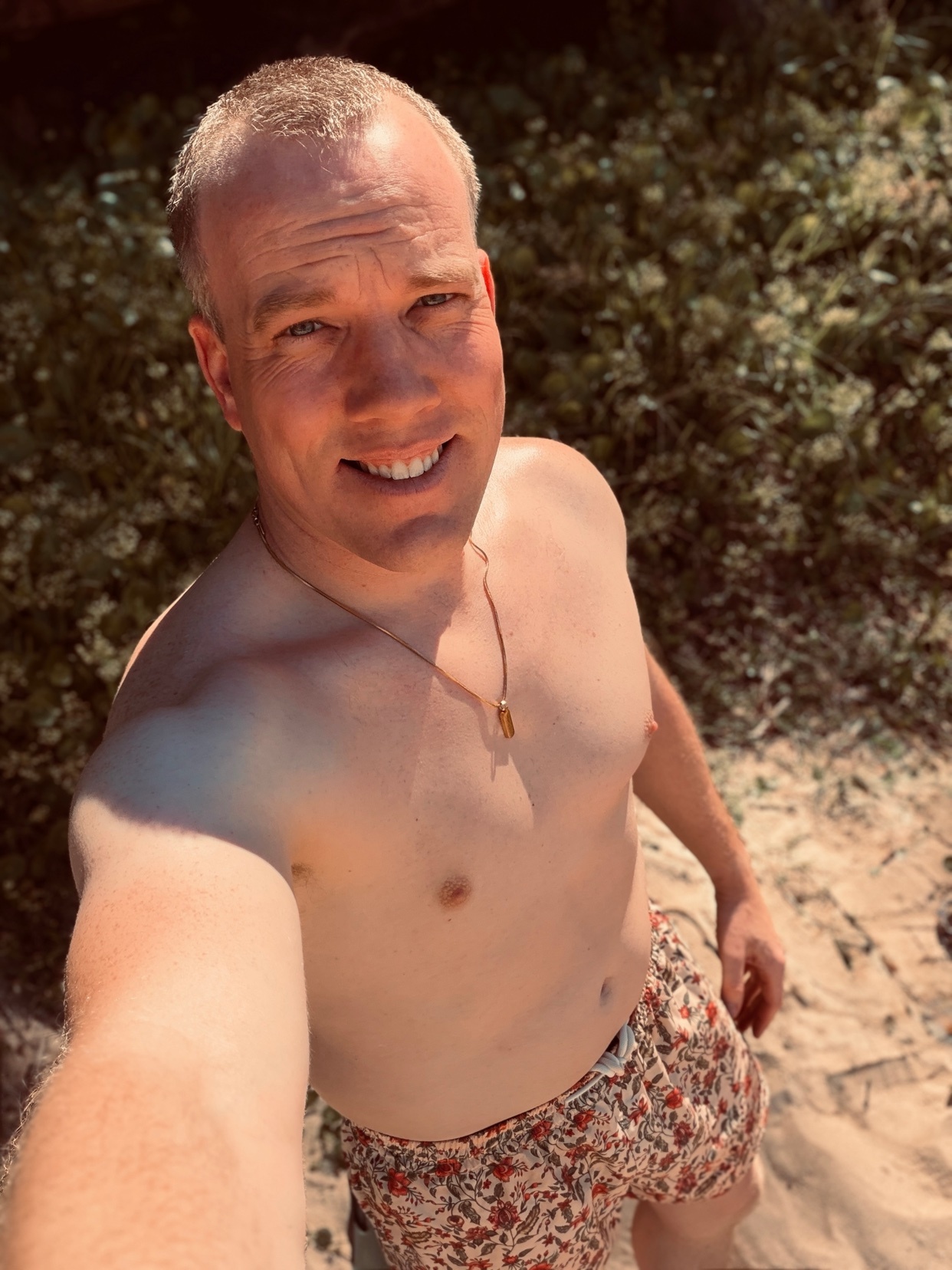 A man in patterned swim trunks smiles at the camera while taking a selfie on a sunny beach. He is shirtless, showing his bare chest and shoulders.