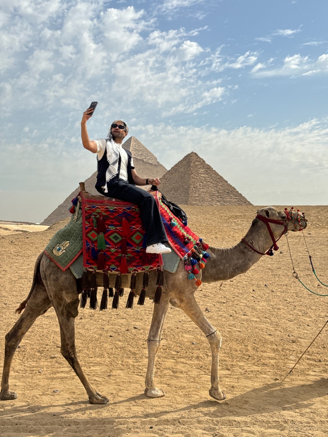 A man takes a selfie while riding a camel in front of the Giza Pyramids under a bright, cloudy sky.