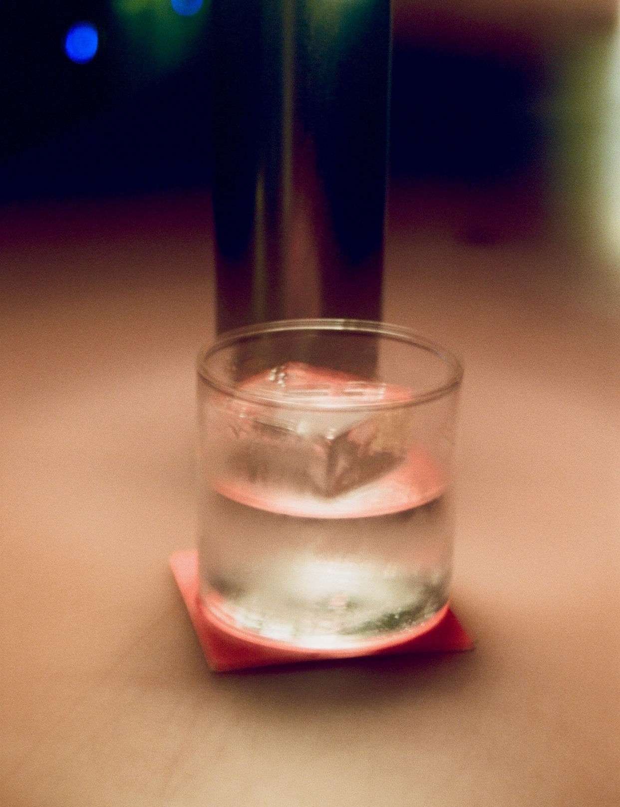 A close-up shot of a clear glass with a large ice cube and a clear liquid, resting on a pink coaster. The background is dark and blurred, with hints of colorful lights.