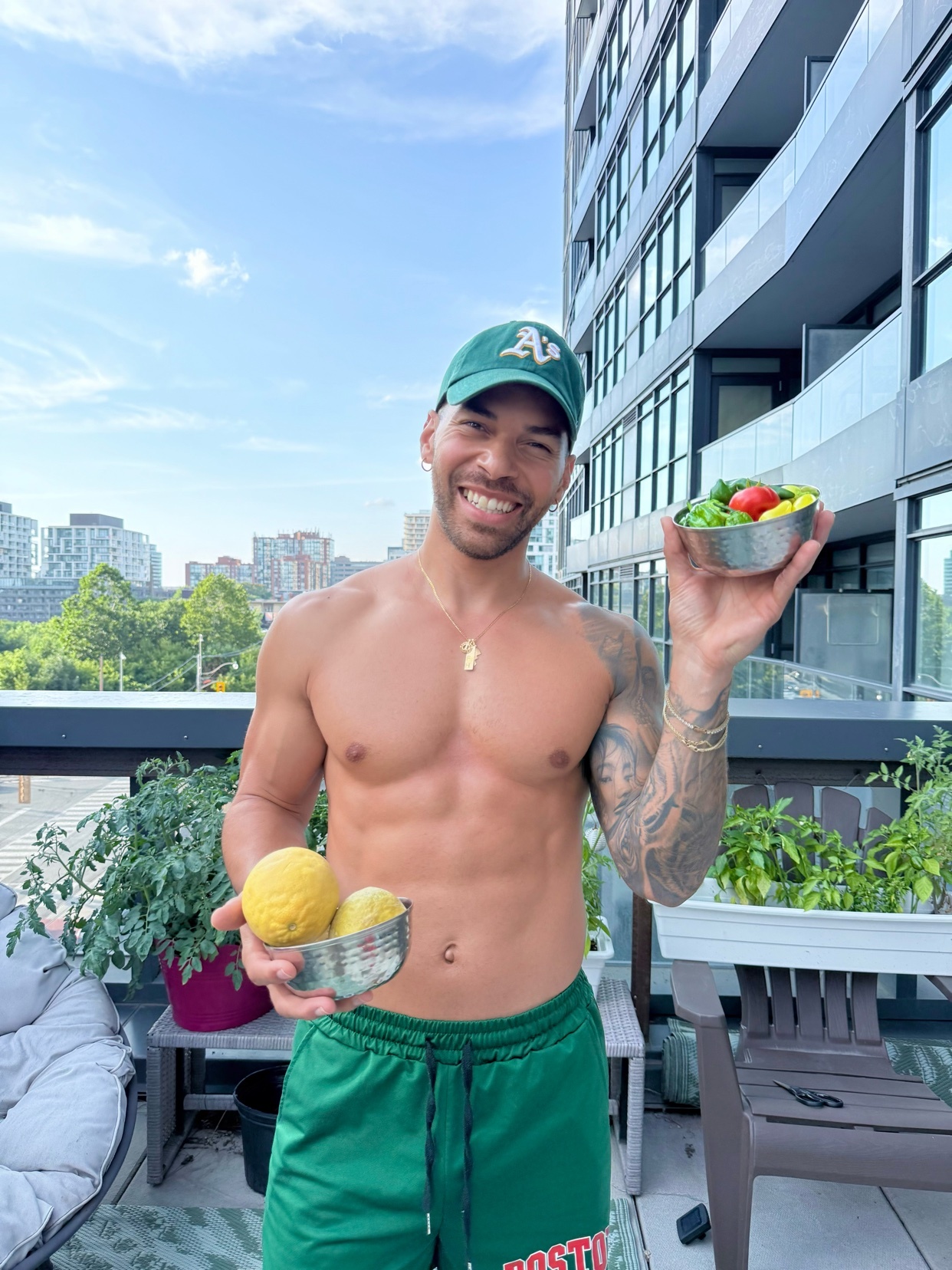 A shirtless man with tattoos, wearing a green hat and shorts, smiles while holding lemons and a small salad on a balcony with a cityscape in the background.