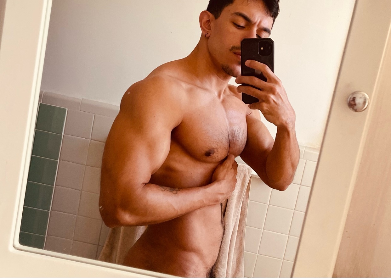A man takes a mirror selfie, showcasing his muscular physique. He is shirtless, highlighting his chest hair and defined muscles.