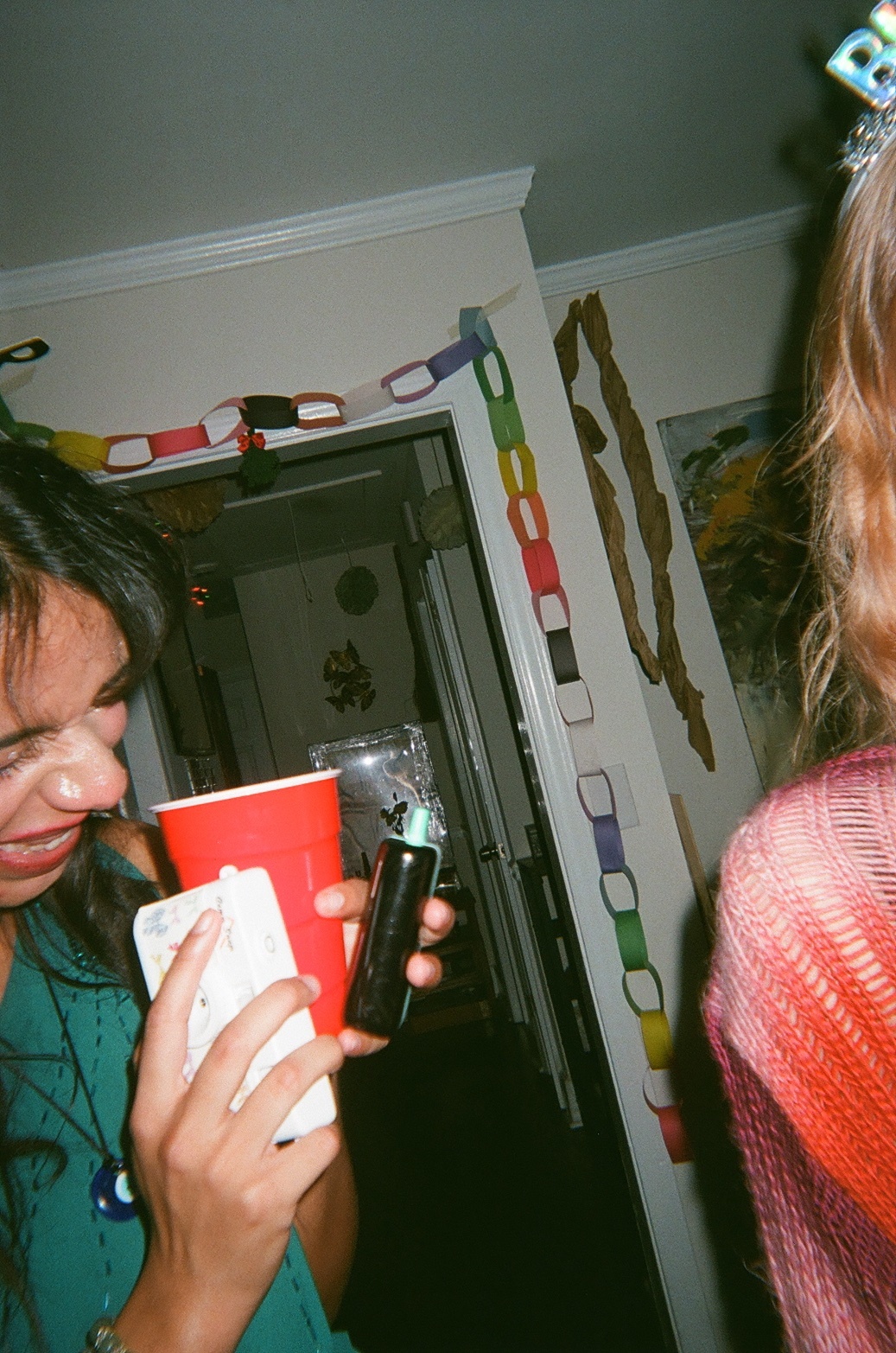 A woman holds a red cup and a phone at a party. A paper chain decorates the doorway.