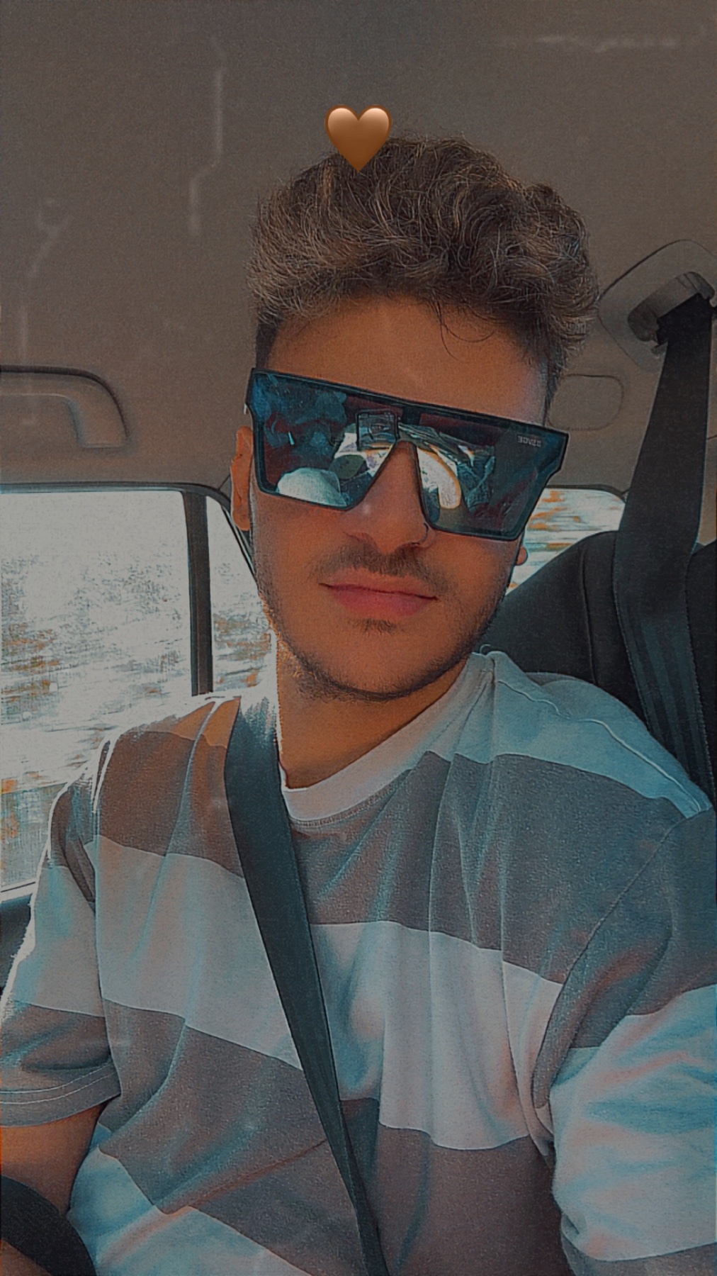 A young man with stylish sunglasses and a striped t-shirt takes a selfie in a car. A small heart emoji floats above his head.