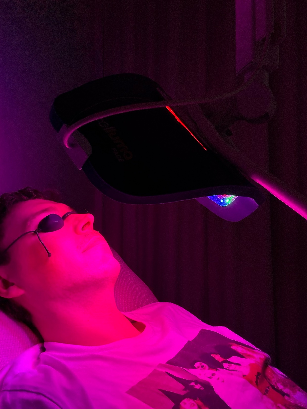 A man lies on his back wearing protective eyewear while undergoing a red light therapy facial treatment. The room is bathed in a vibrant pink and purple glow from the LED light machine.