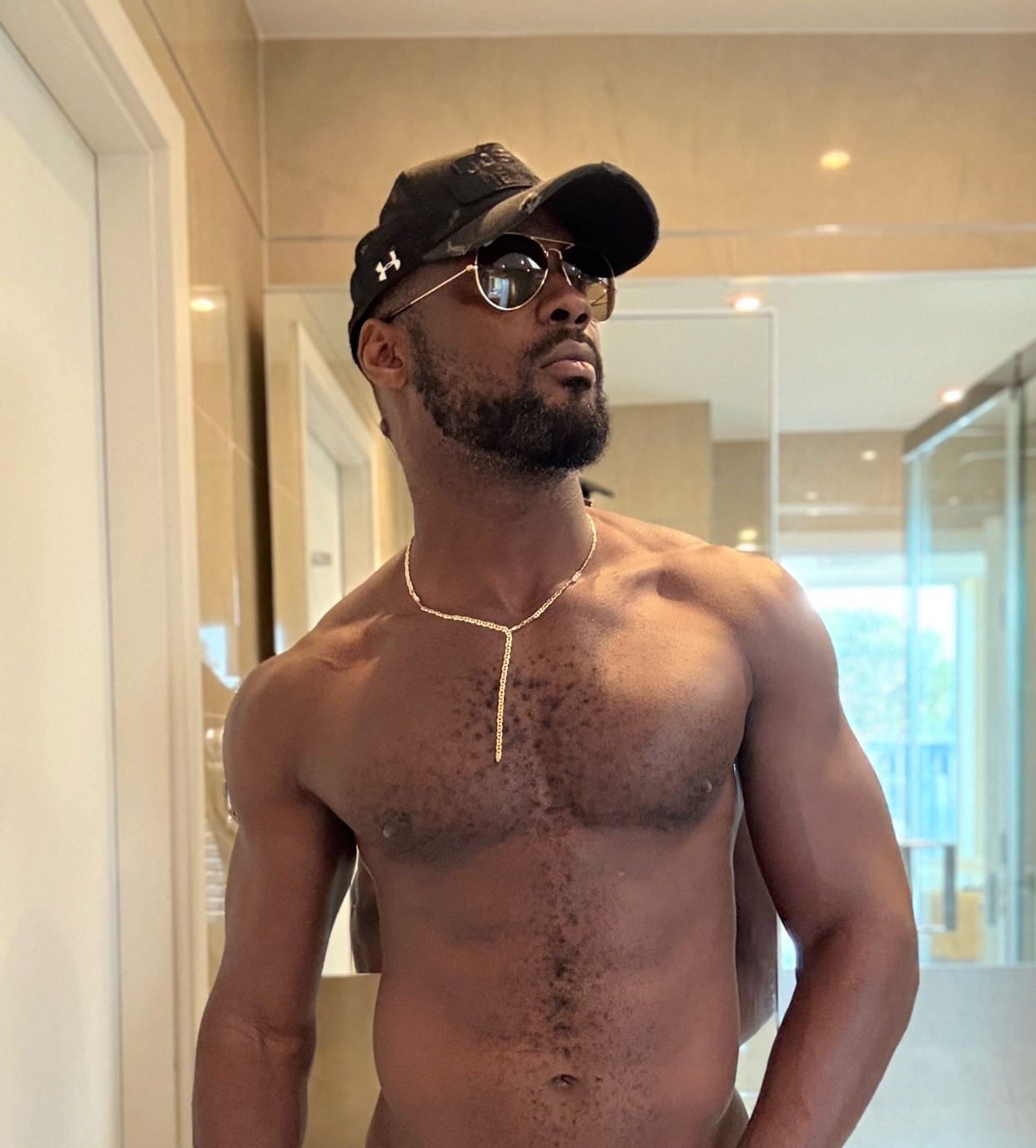 A shirtless man poses in a bathroom, wearing a cap and sunglasses. He has a muscular physique and a gold necklace.