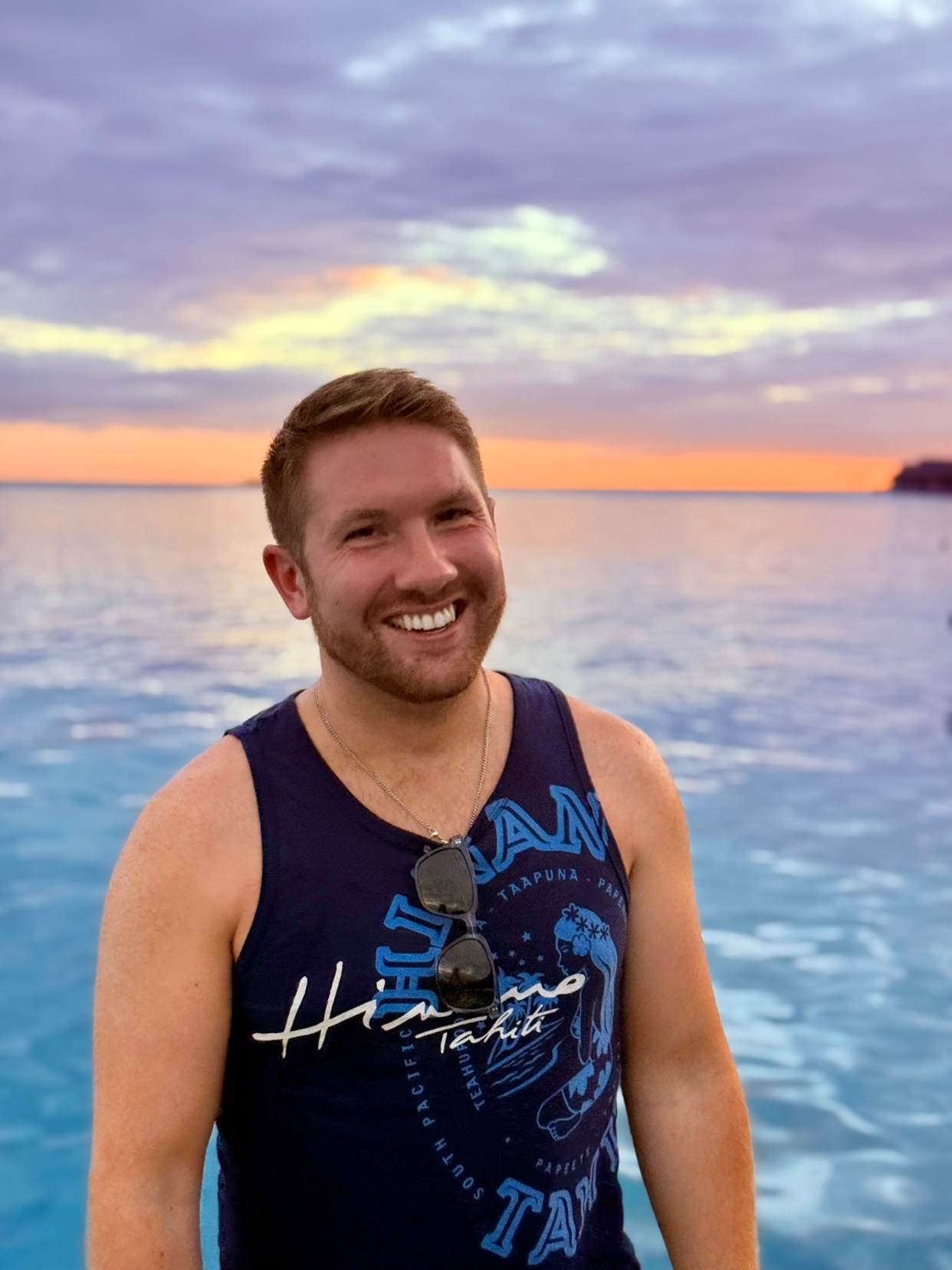 A man smiles at the camera wearing a dark blue tank top and sunglasses perched on his head. He stands in front of a bright blue ocean with a colorful sunset sky in the background.