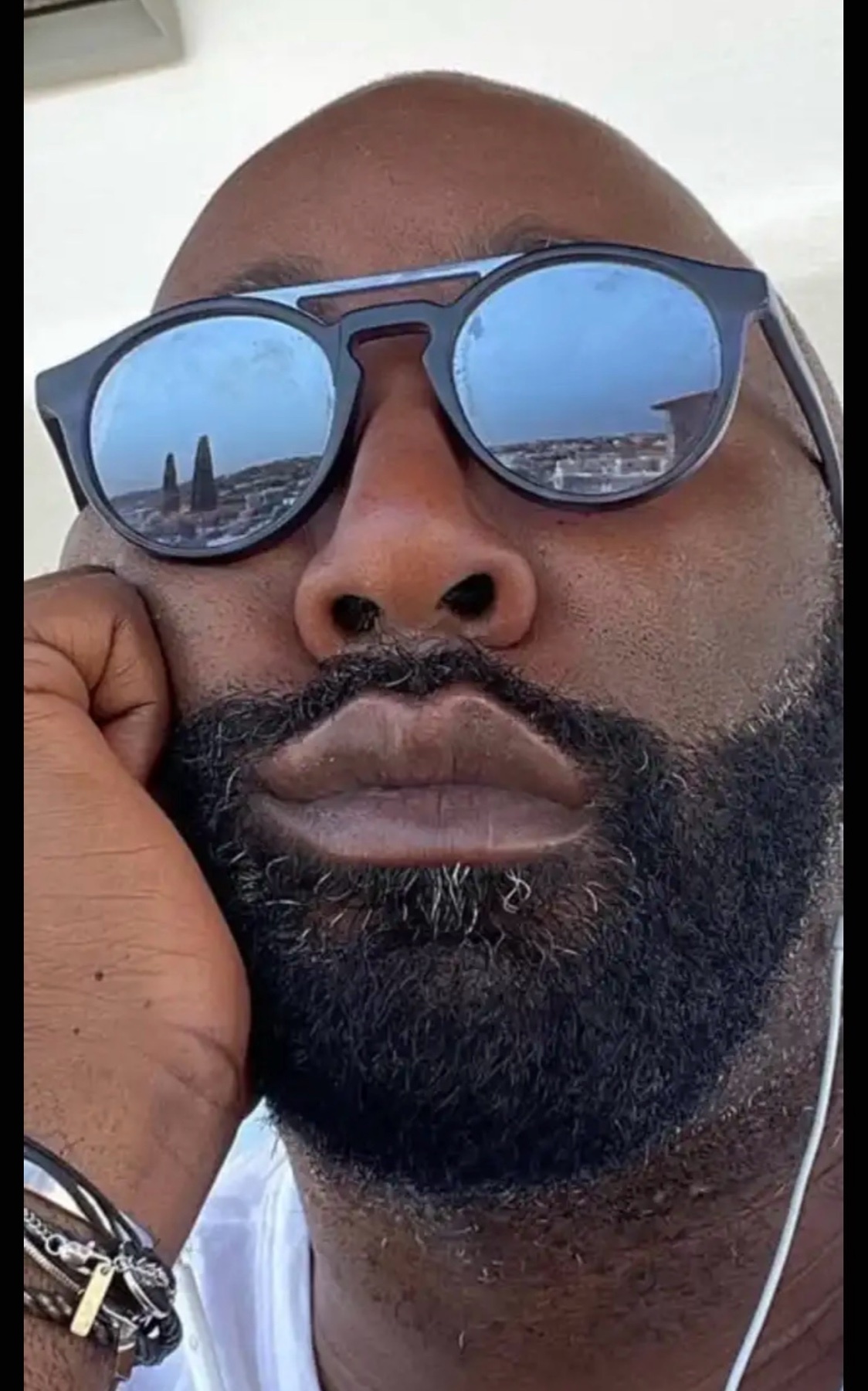 A close-up shot of a bald Black man wearing sunglasses and a beard. The sunglasses reflect a cityscape with trees.