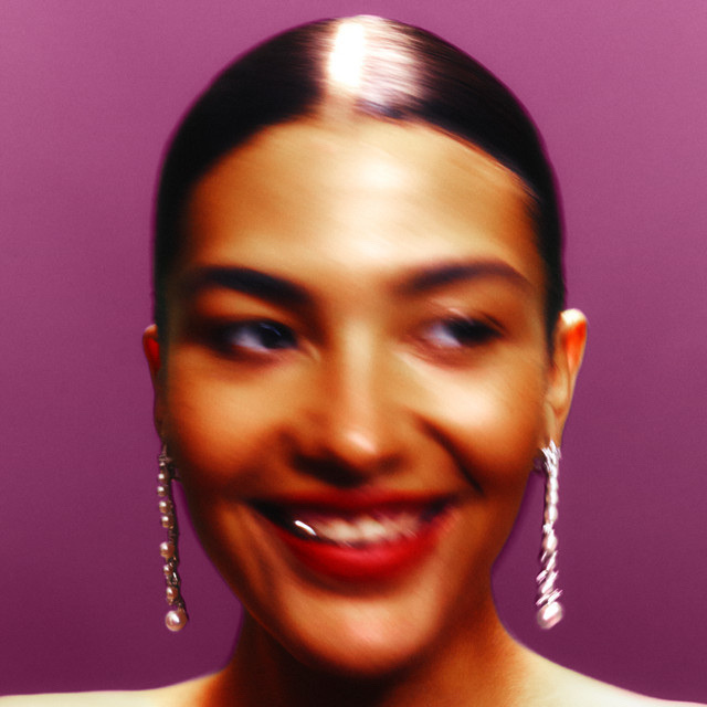 A woman with dark hair and earrings smiles in a portrait. The image has a soft, blurred effect.