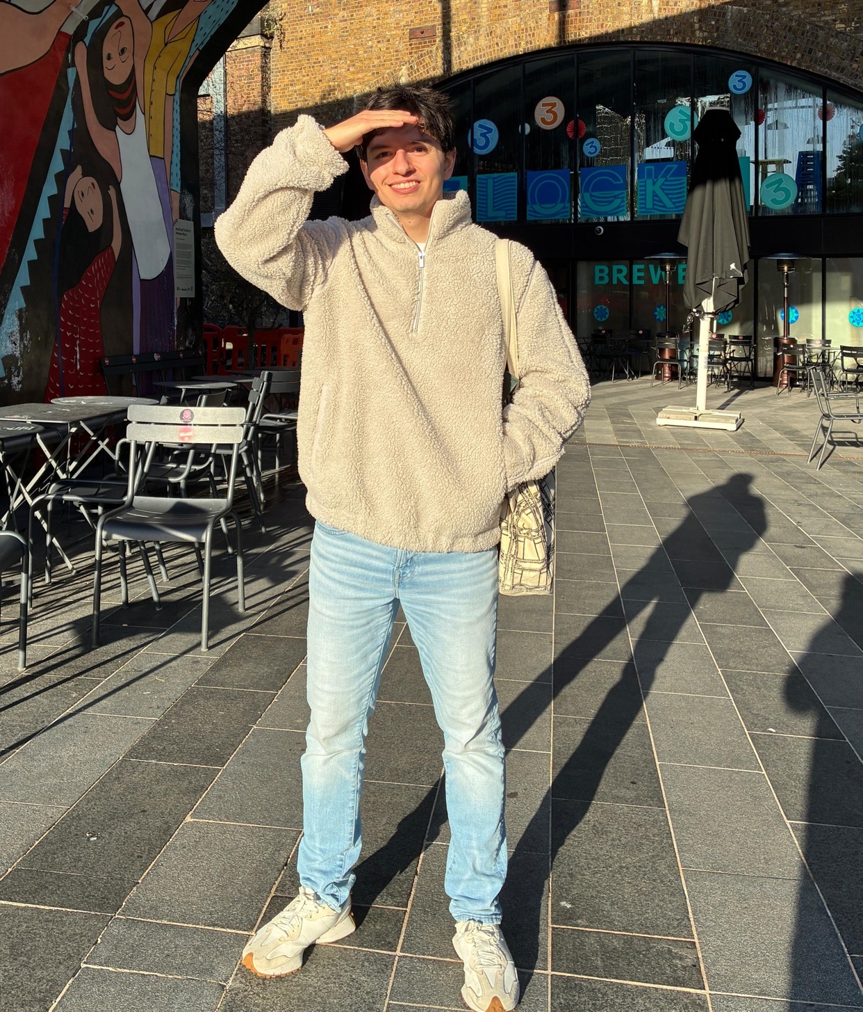 A man poses in a casual outfit, shielding his eyes from the sun. He is wearing jeans, a fleece, and sneakers.