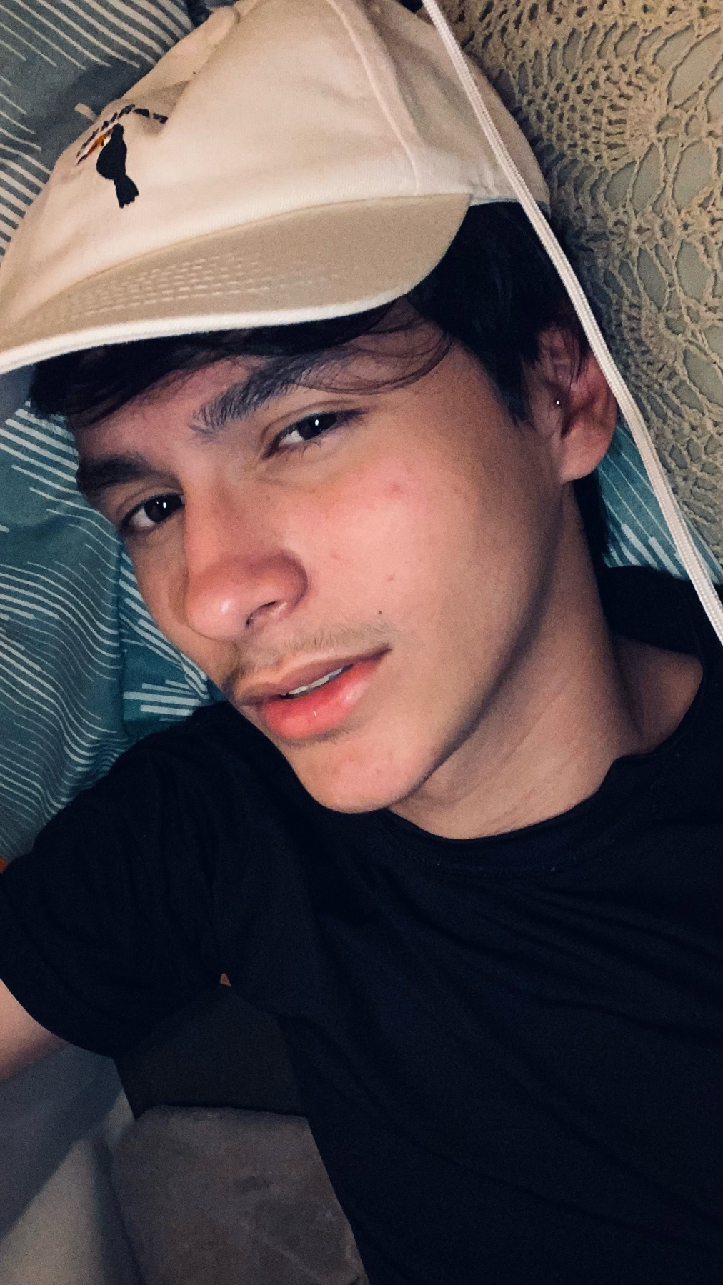 A young man wearing a white baseball cap and a black t-shirt poses for a selfie. He has dark hair and light stubble on his face.