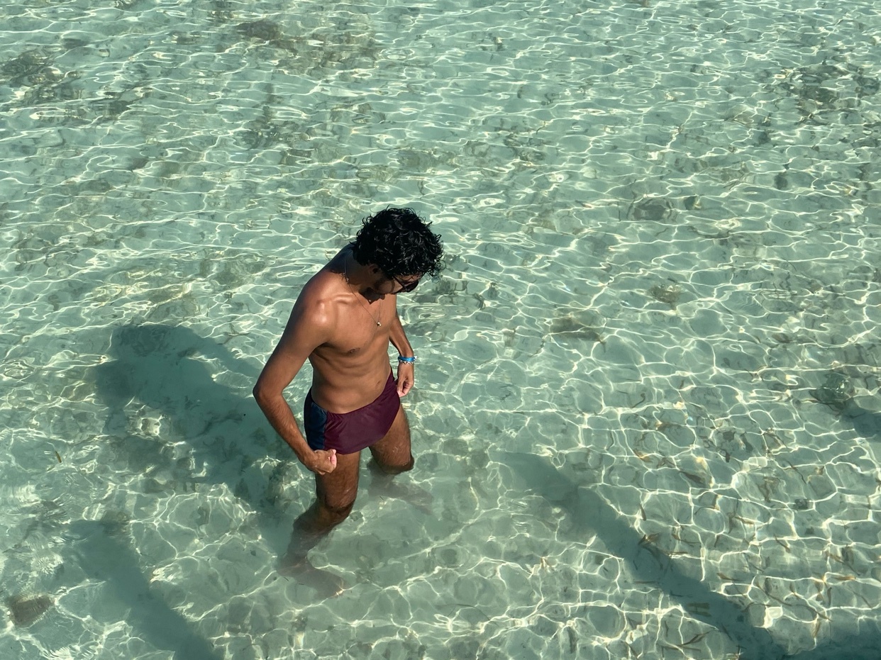 A man stands in crystal clear turquoise water wearing maroon swim trunks and sunglasses. The sunlight creates a beautiful dappled pattern on the sandy ocean floor beneath him.