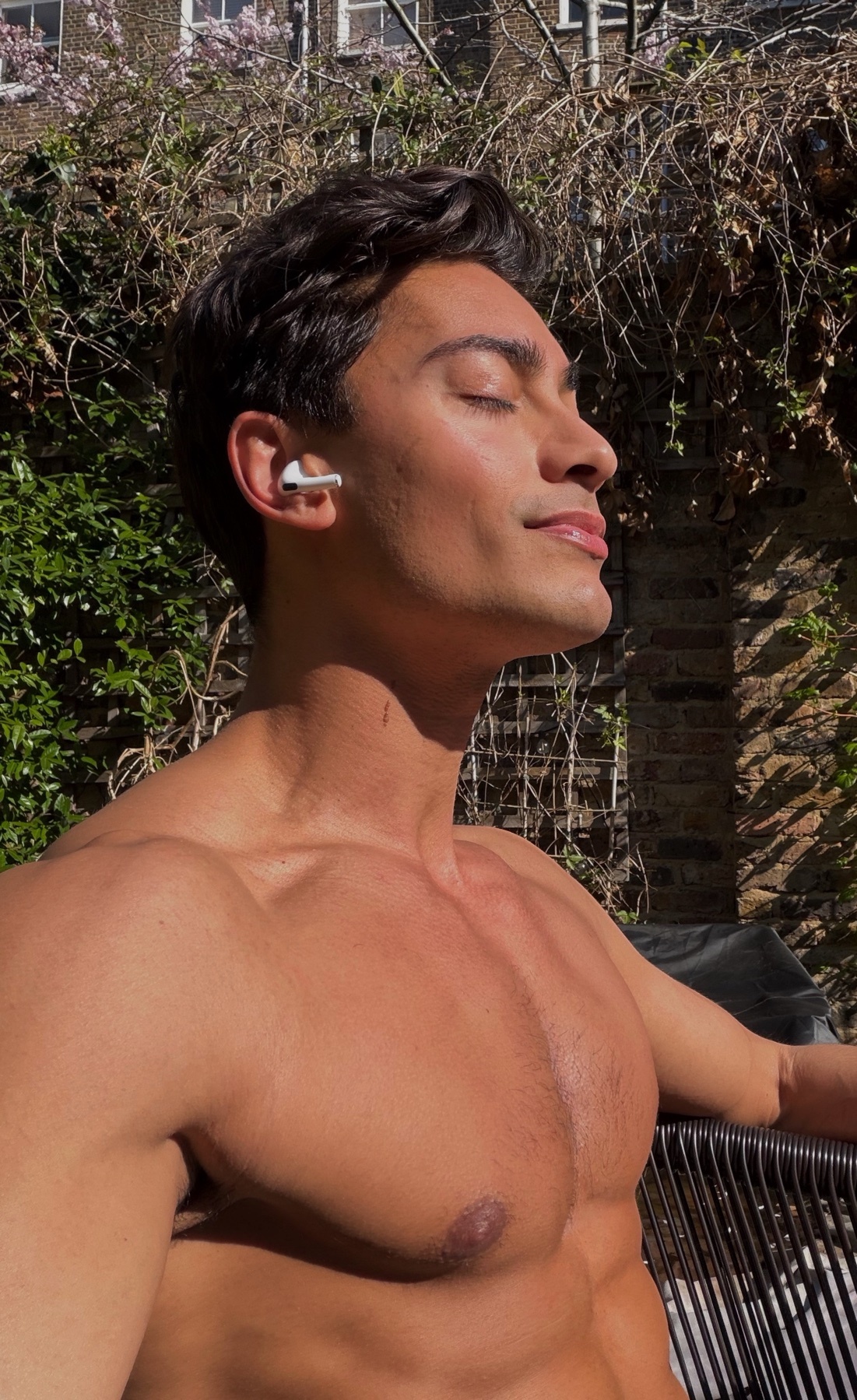 A shirtless man with dark hair and earbuds is basking in the sun, eyes closed, with a relaxed expression. He is outdoors with greenery and brickwork in the background.
