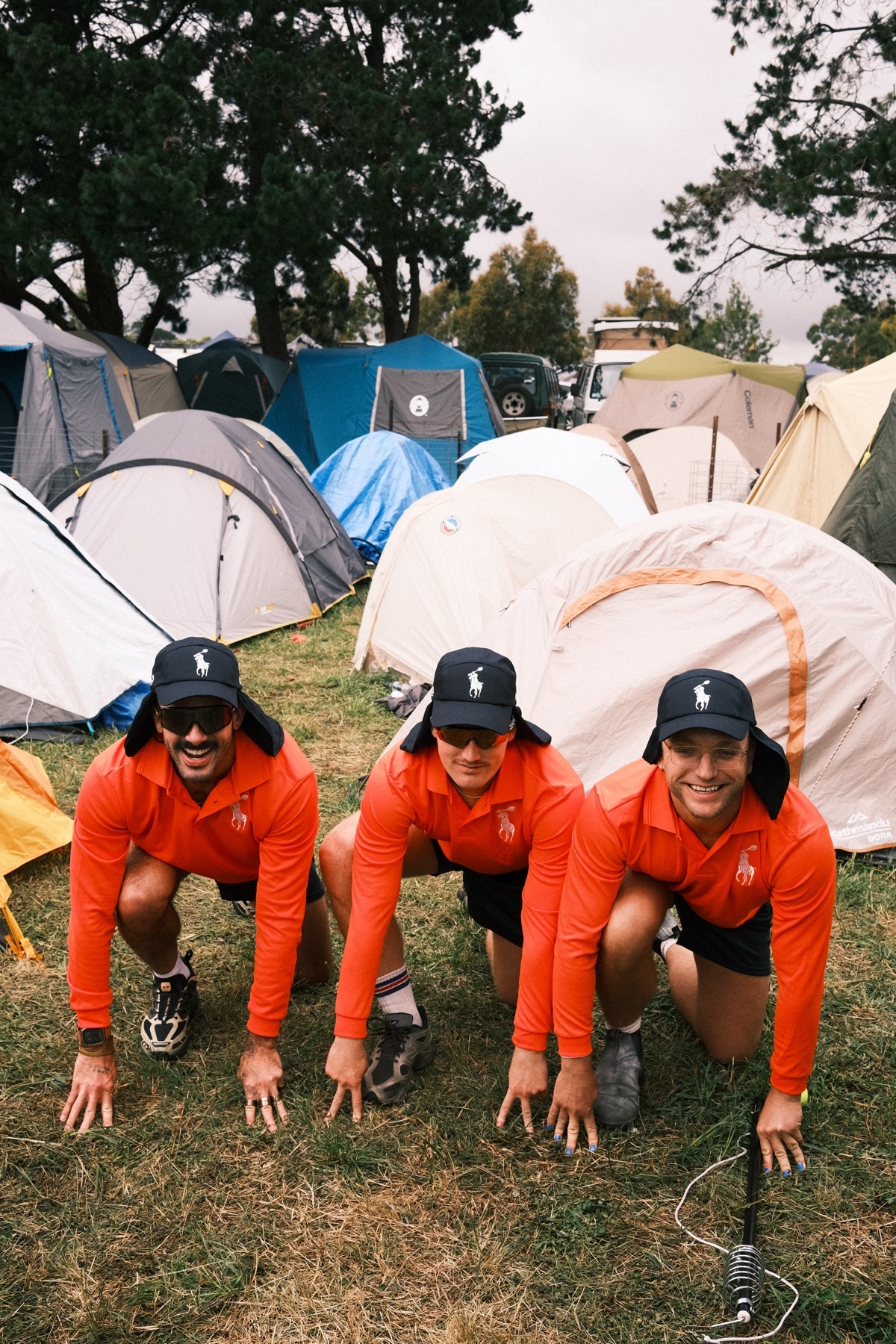 Three men in matching orange shirts and black shorts kneel on a grassy field, posing for a photo. They wear black polo hats and appear to be ball boys.