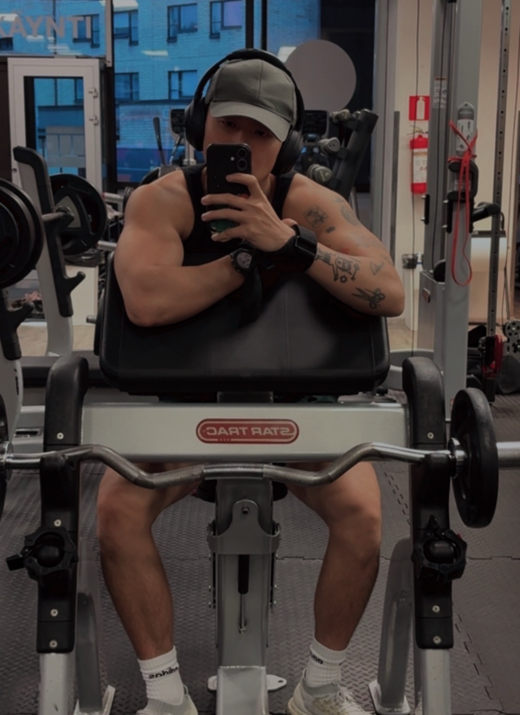 A man takes a selfie in a gym while sitting on a weightlifting machine. He is wearing headphones and a baseball cap, and has tattoos on his arms.