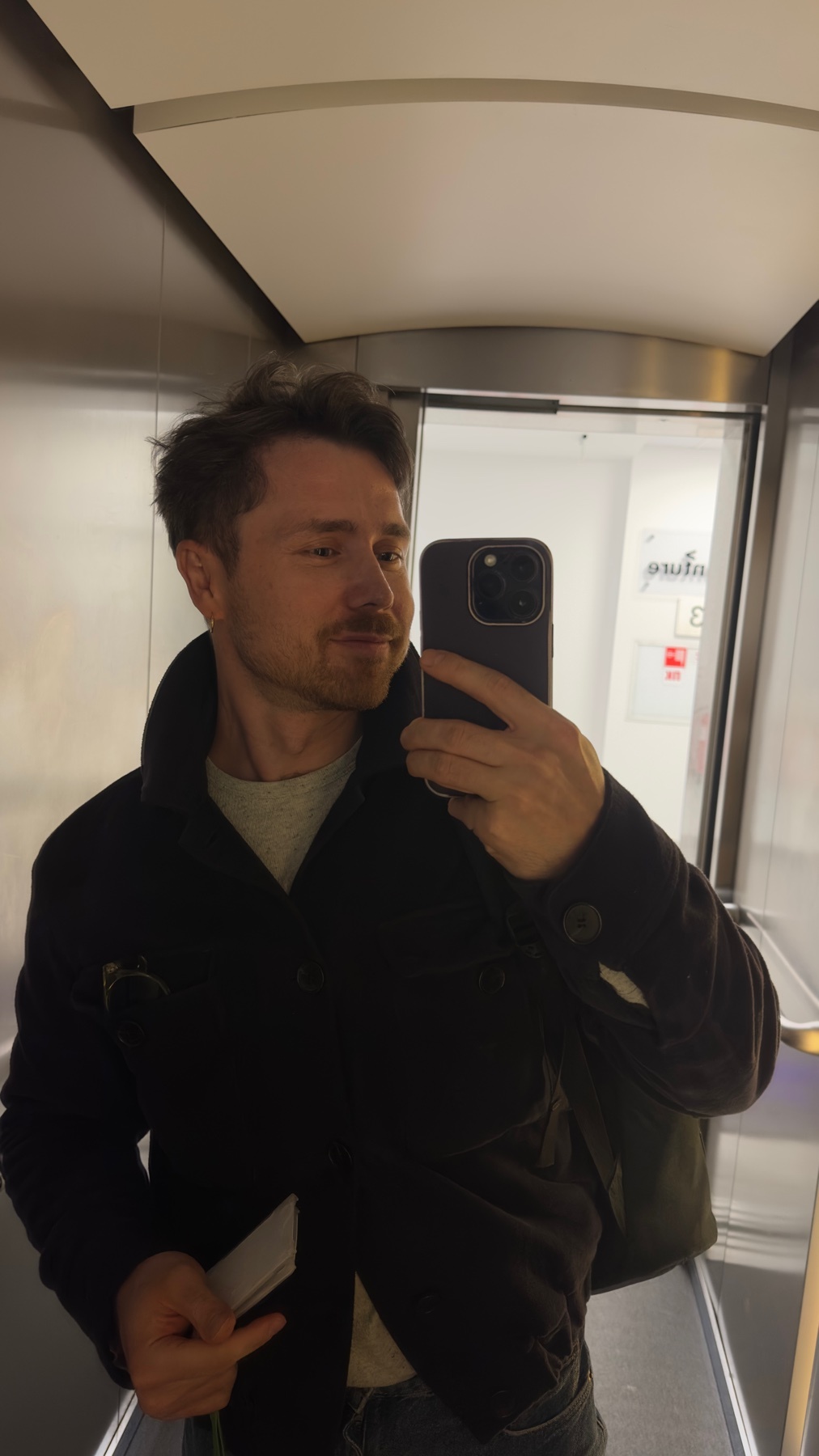 A man takes a selfie in an elevator, wearing a dark jacket over a grey t-shirt. He has short, dark hair and a beard, and is looking at his phone.