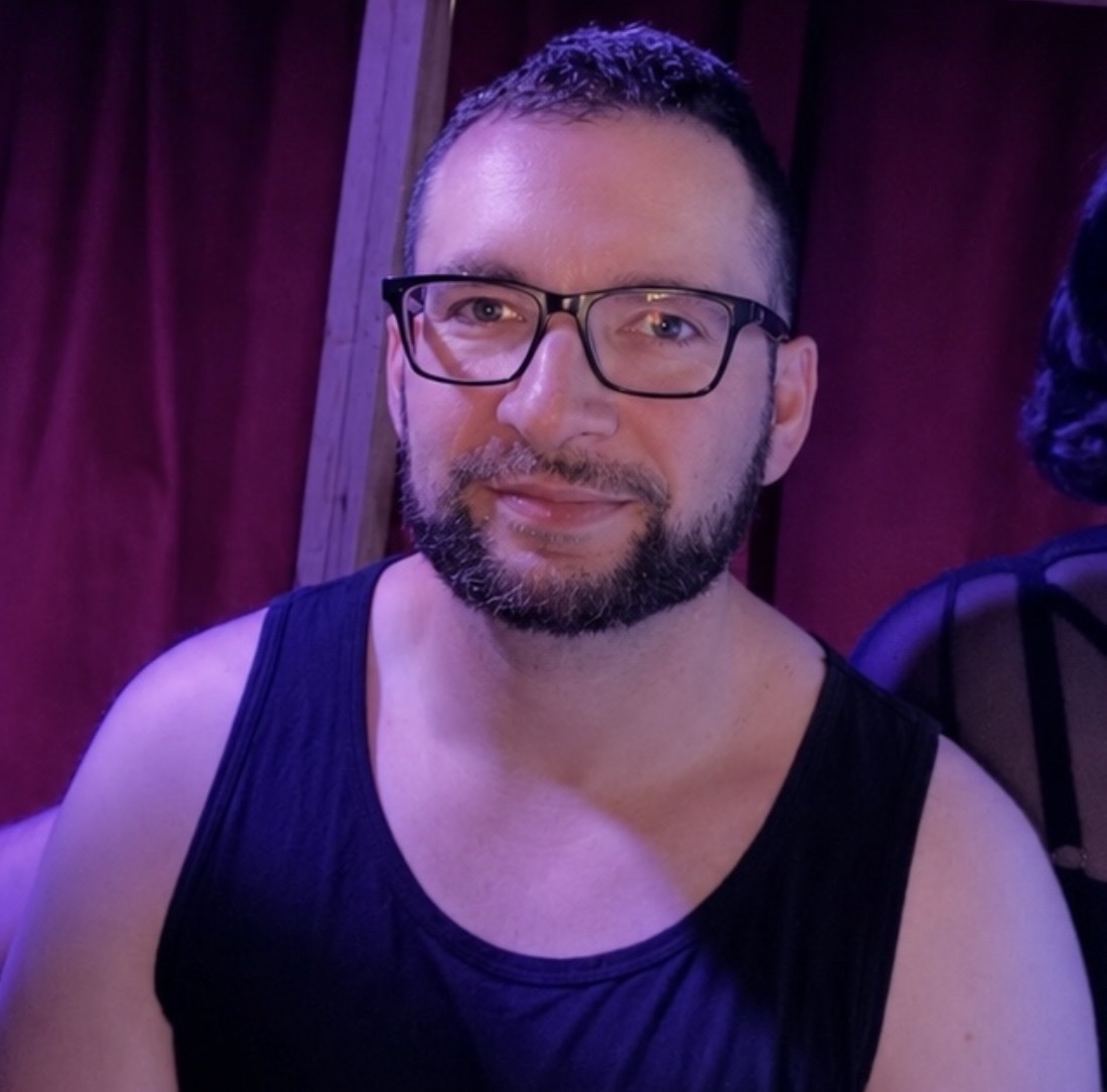 A man with a beard and glasses smiles at the camera while wearing a black tank top.