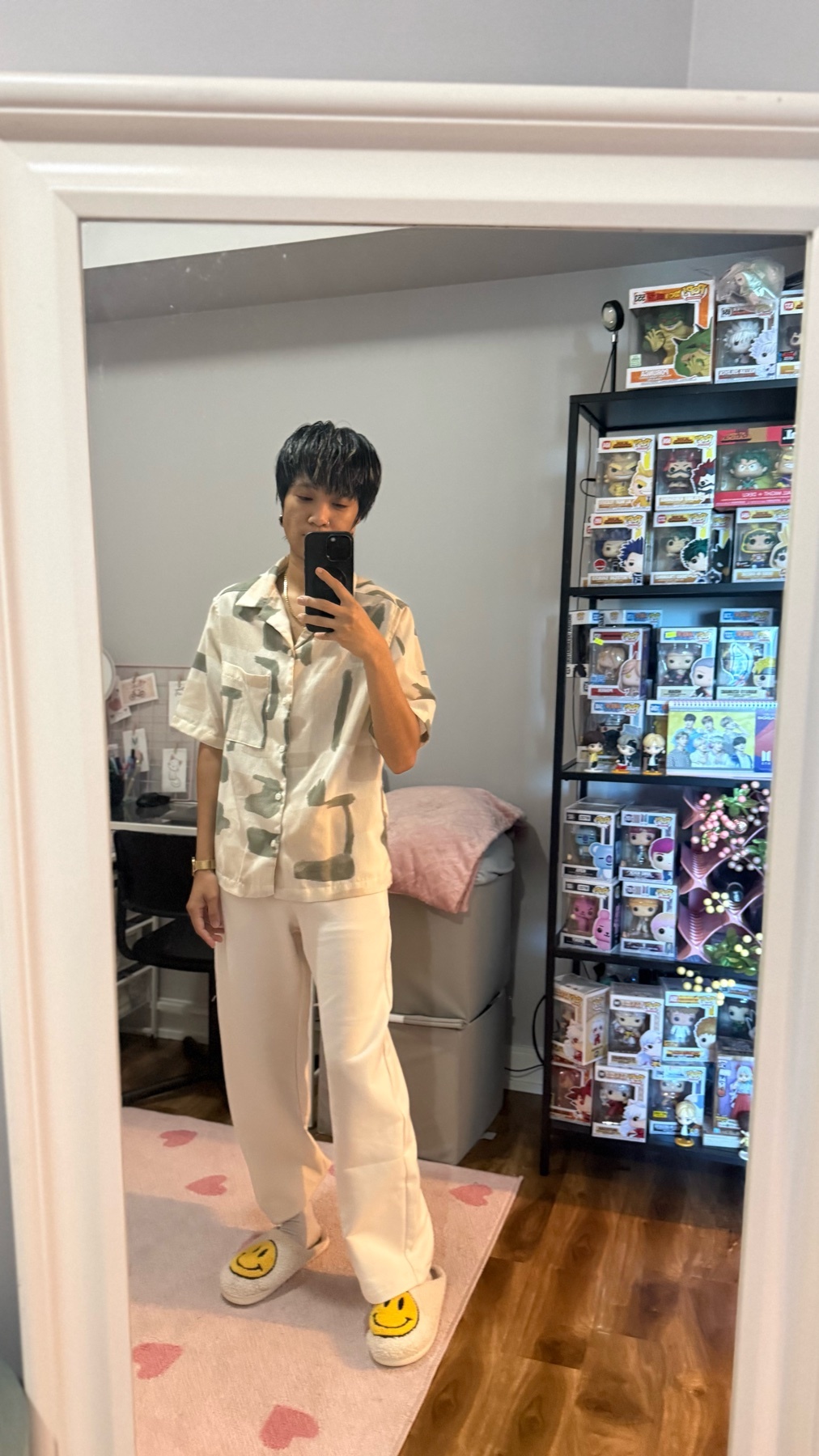A non-binary person stands in front of a mirror, taking a selfie. They are wearing a satin button-up shirt with a camouflage pattern, loose-fitting cream pants, and yellow clogs with smiley faces on them. The background shows a shelf filled with coll
