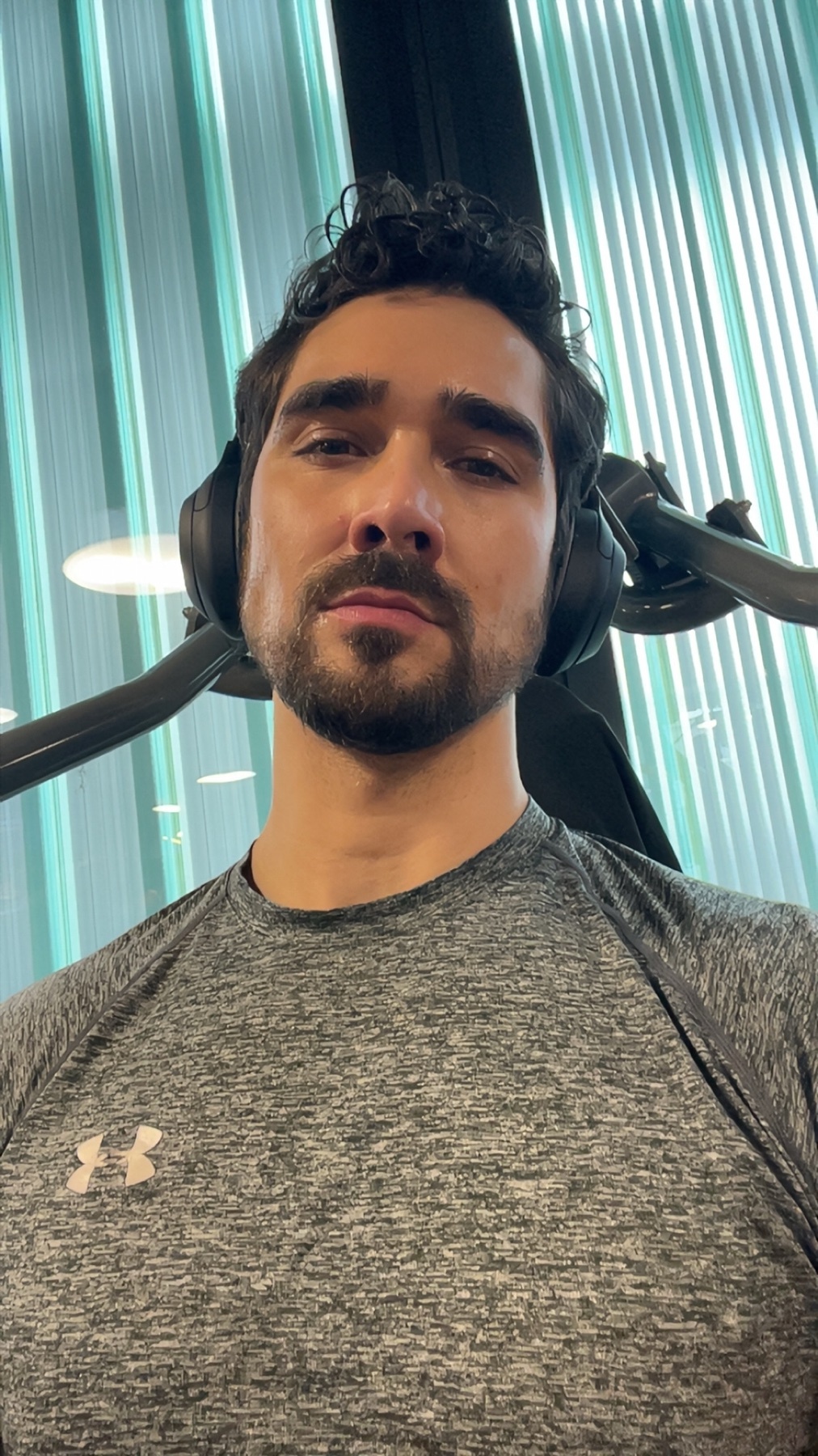 A man with dark, curly hair and stubble is wearing headphones and a grey t-shirt. He appears to be at the gym, possibly taking a selfie.