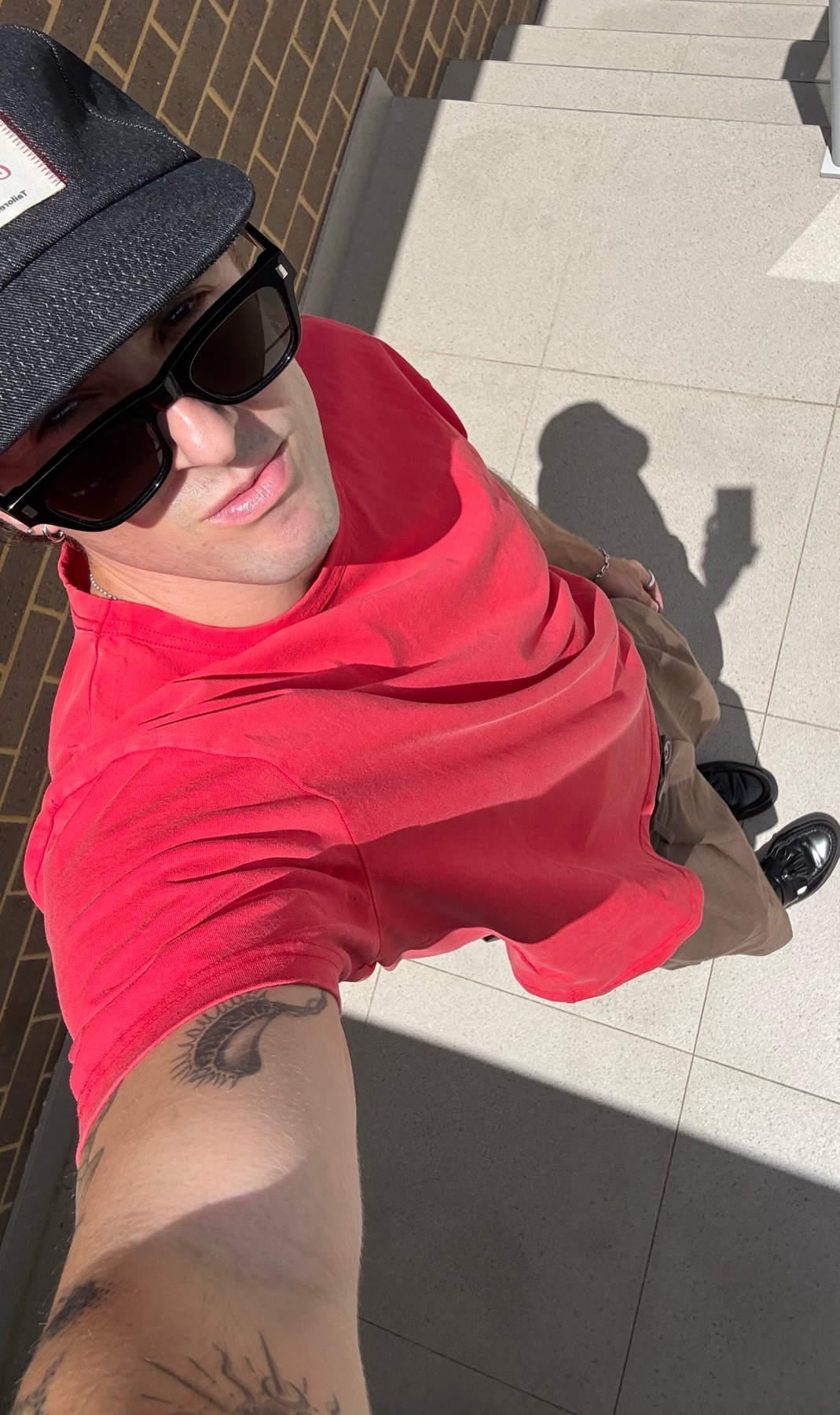 A man takes a selfie in the sun, wearing a red t-shirt, sunglasses, and a cap. He has a tattoo on his arm.