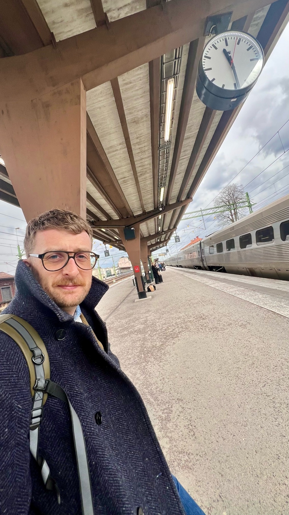 A man with glasses and a mustache takes a selfie on a train station platform. He is wearing a dark coat and a backpack, with a train visible in the background.
