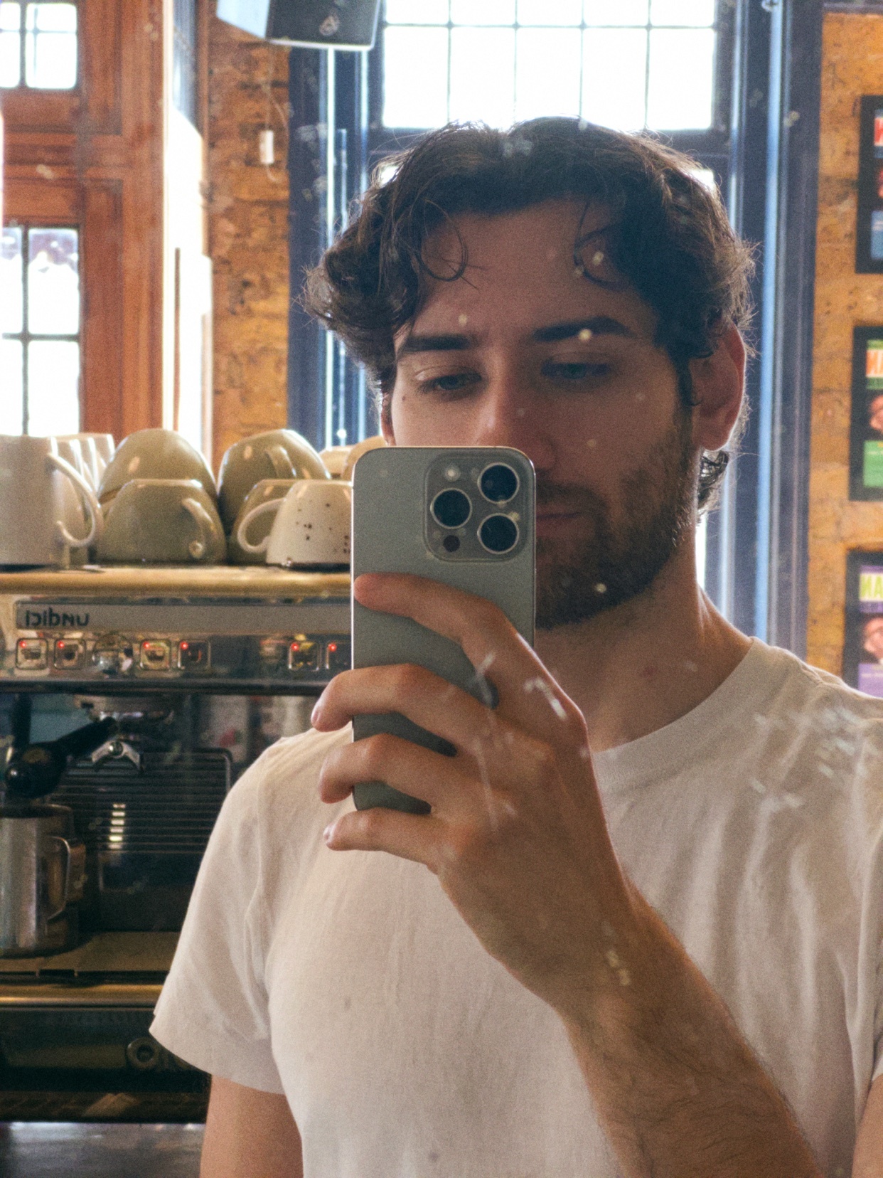 A man with dark, curly hair and stubble takes a selfie in a coffee shop. He is wearing a white t-shirt and holding a smartphone.