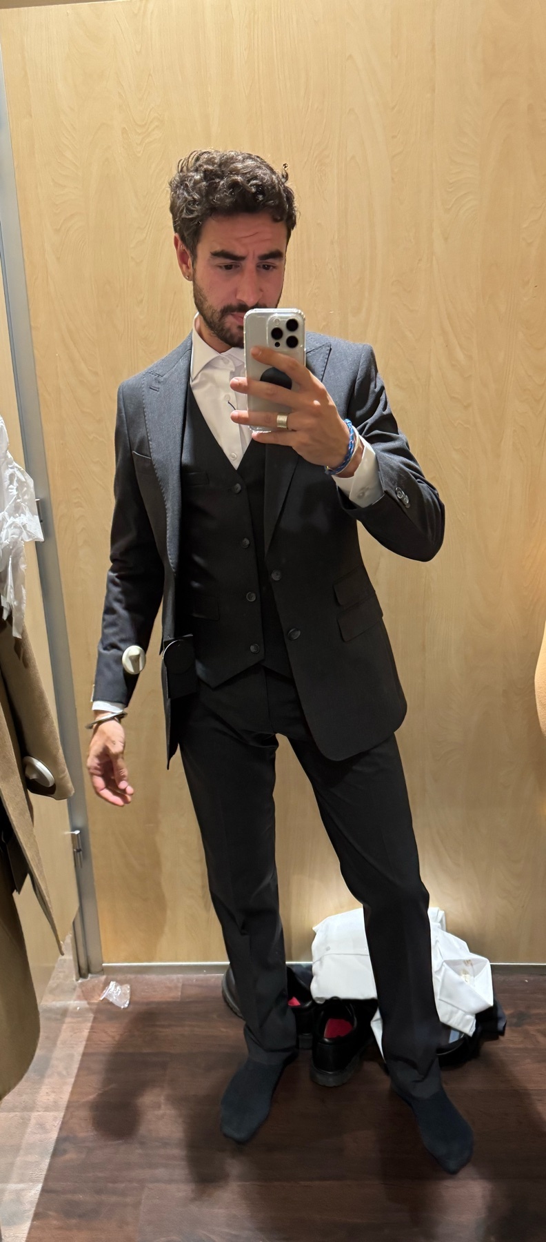 A man in a dark grey three-piece suit and white shirt poses for a mirror selfie in a dressing room. He is wearing a suit jacket, vest, and trousers, with black dress shoes.