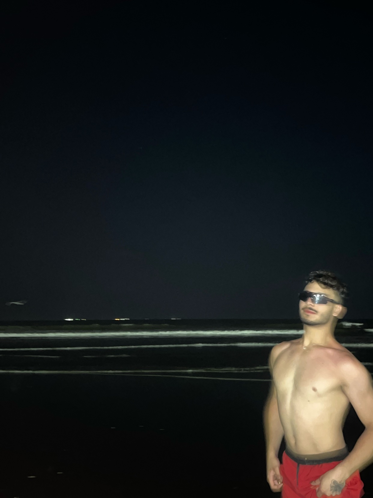 A shirtless man poses on a beach at night. He is wearing sunglasses and red shorts.
