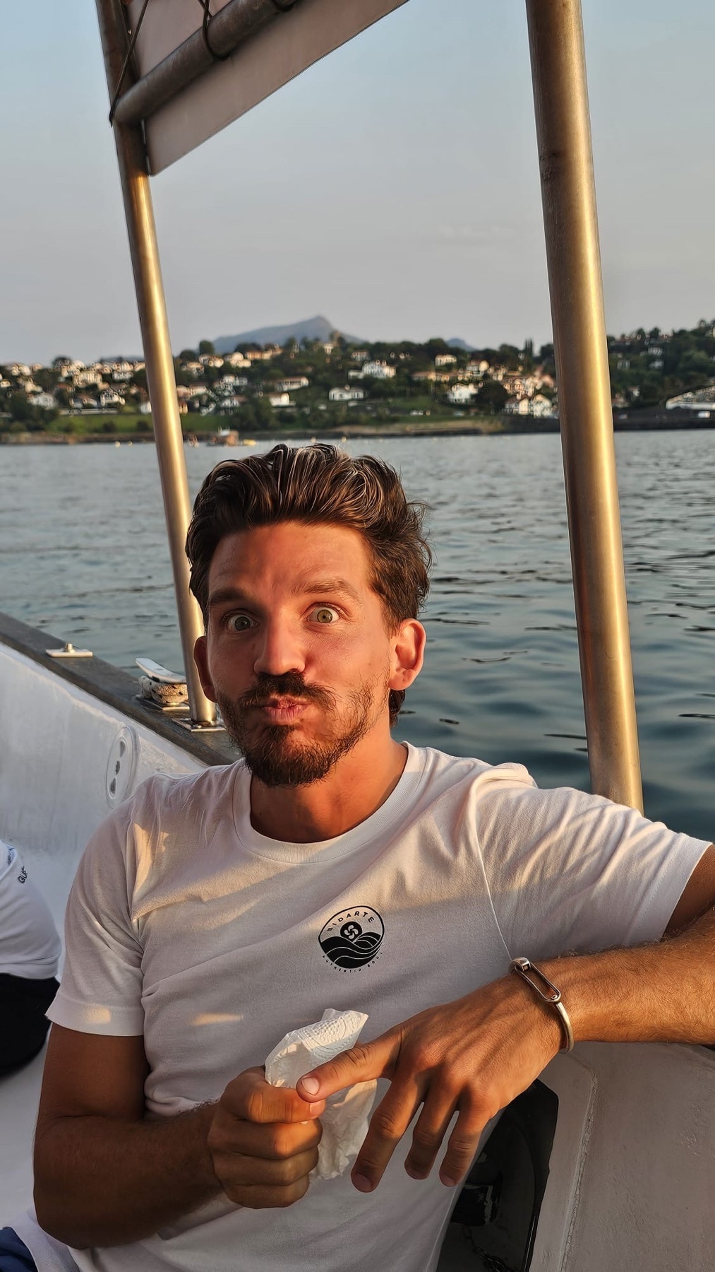 A man makes a funny face while on a boat. The photo is taken during the day with water and a coastline in the background.