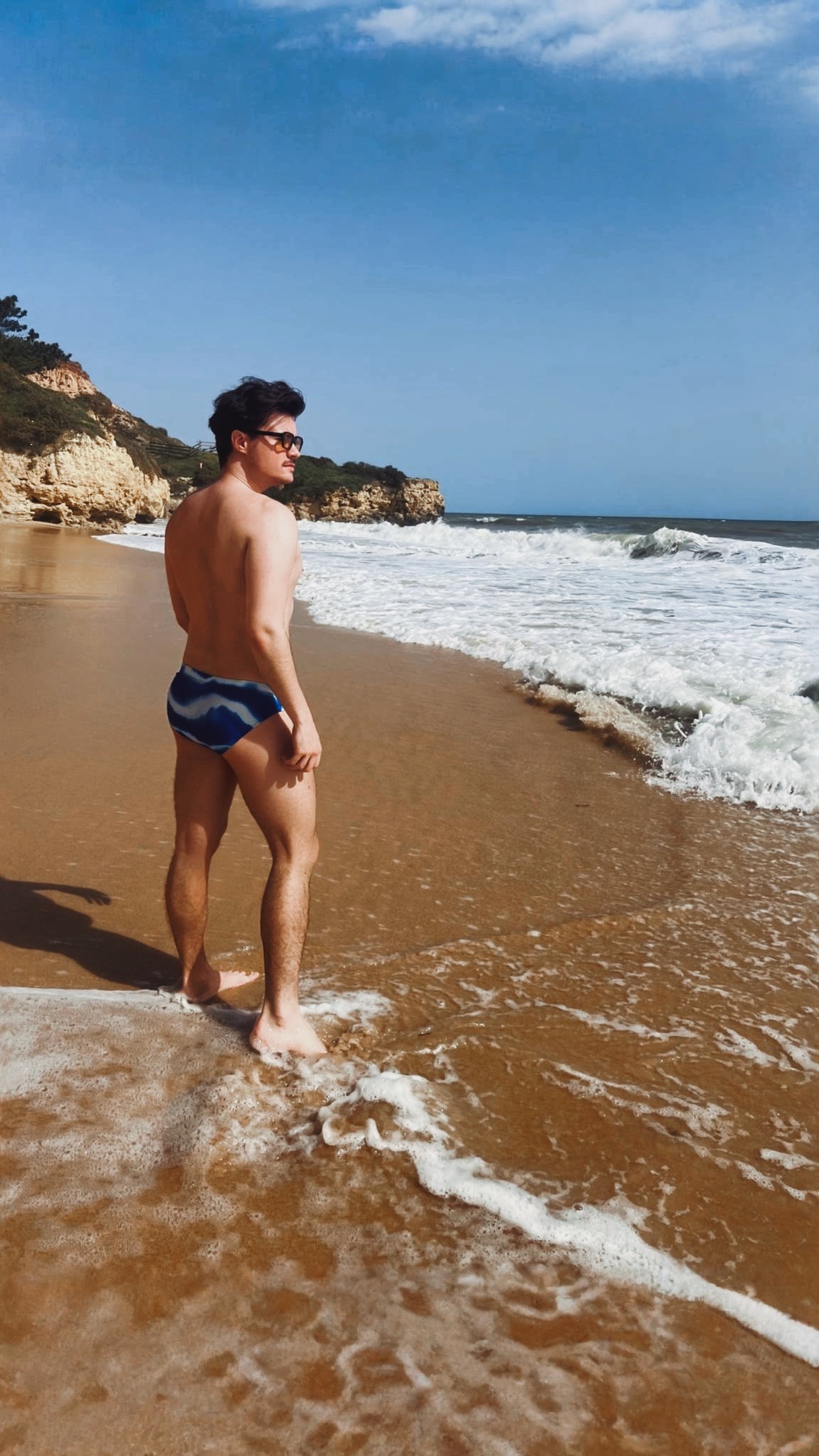 A man in blue and white striped swim trunks stands on a sandy beach, looking out at the ocean waves crashing on the shore. The sun shines brightly on the coastline under a blue sky with scattered clouds.
