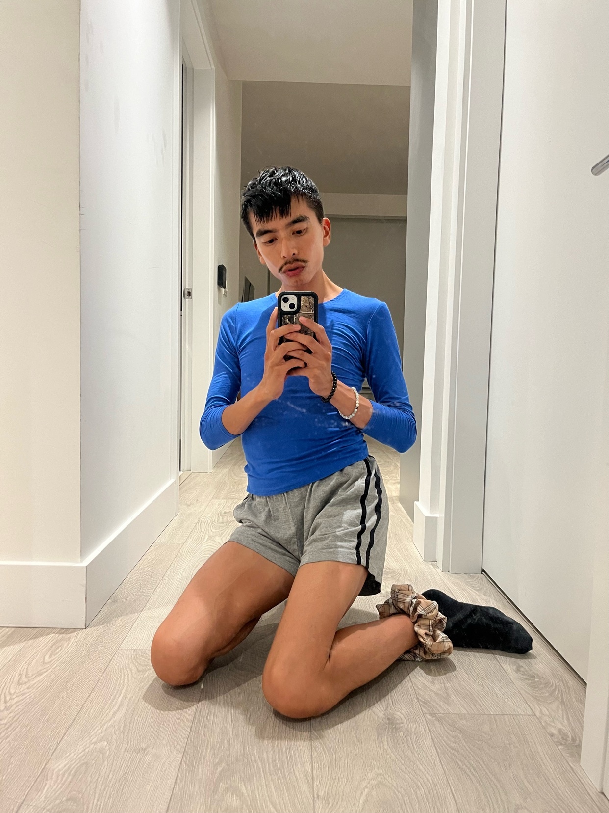 A man kneels on the floor, taking a mirror selfie. He is wearing a blue long-sleeved shirt and grey shorts.