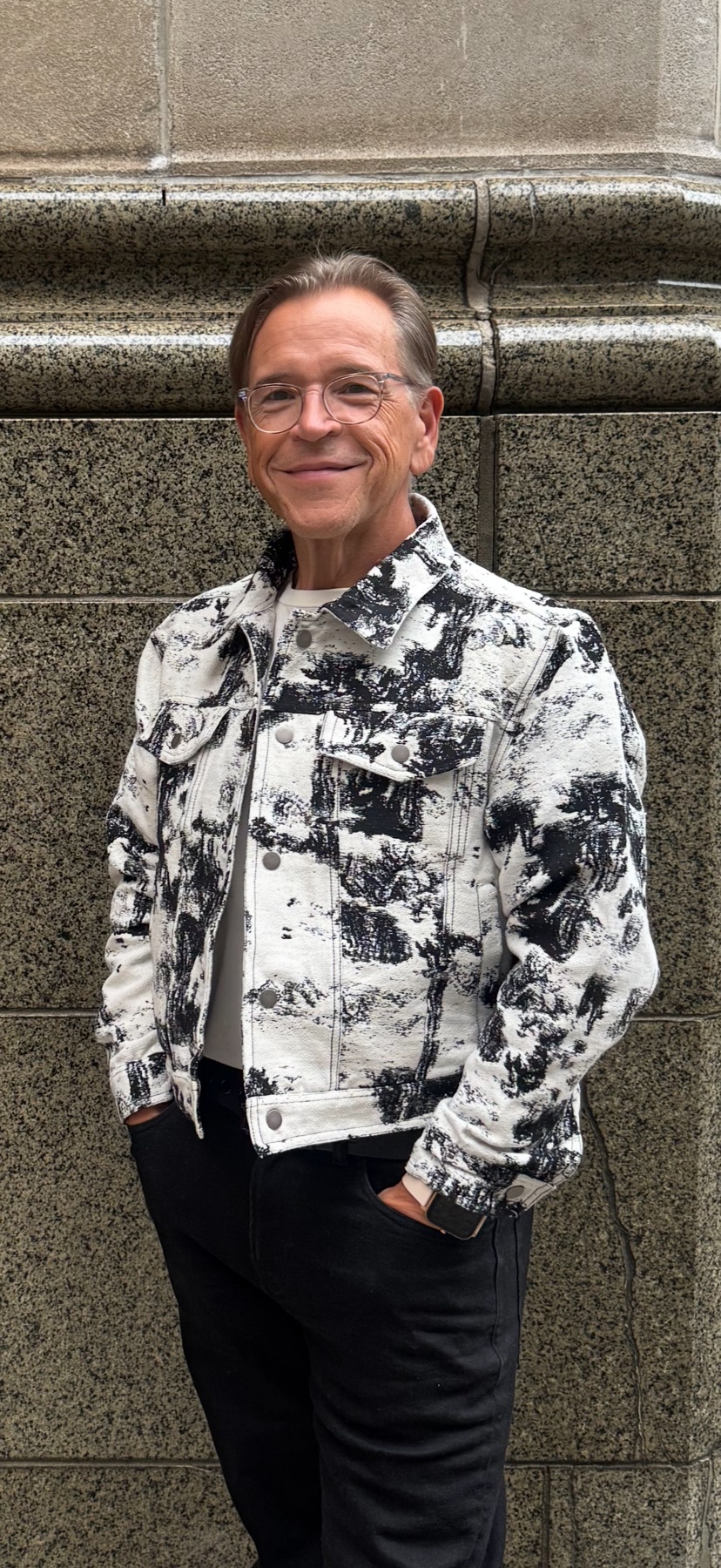 A man with glasses stands in front of a stone wall, wearing a white denim jacket with a black abstract pattern over a white t-shirt and black pants. He has his hands in his pockets and is looking at the camera with a slight smile.