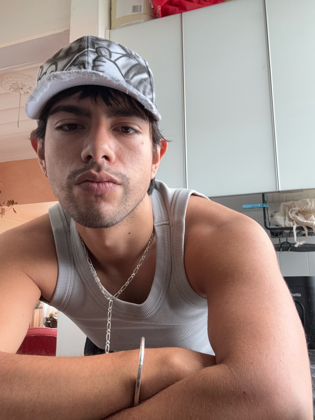 A man takes a selfie in a tank top and cap. He is shirtless and wearing a silver necklace and bracelet.