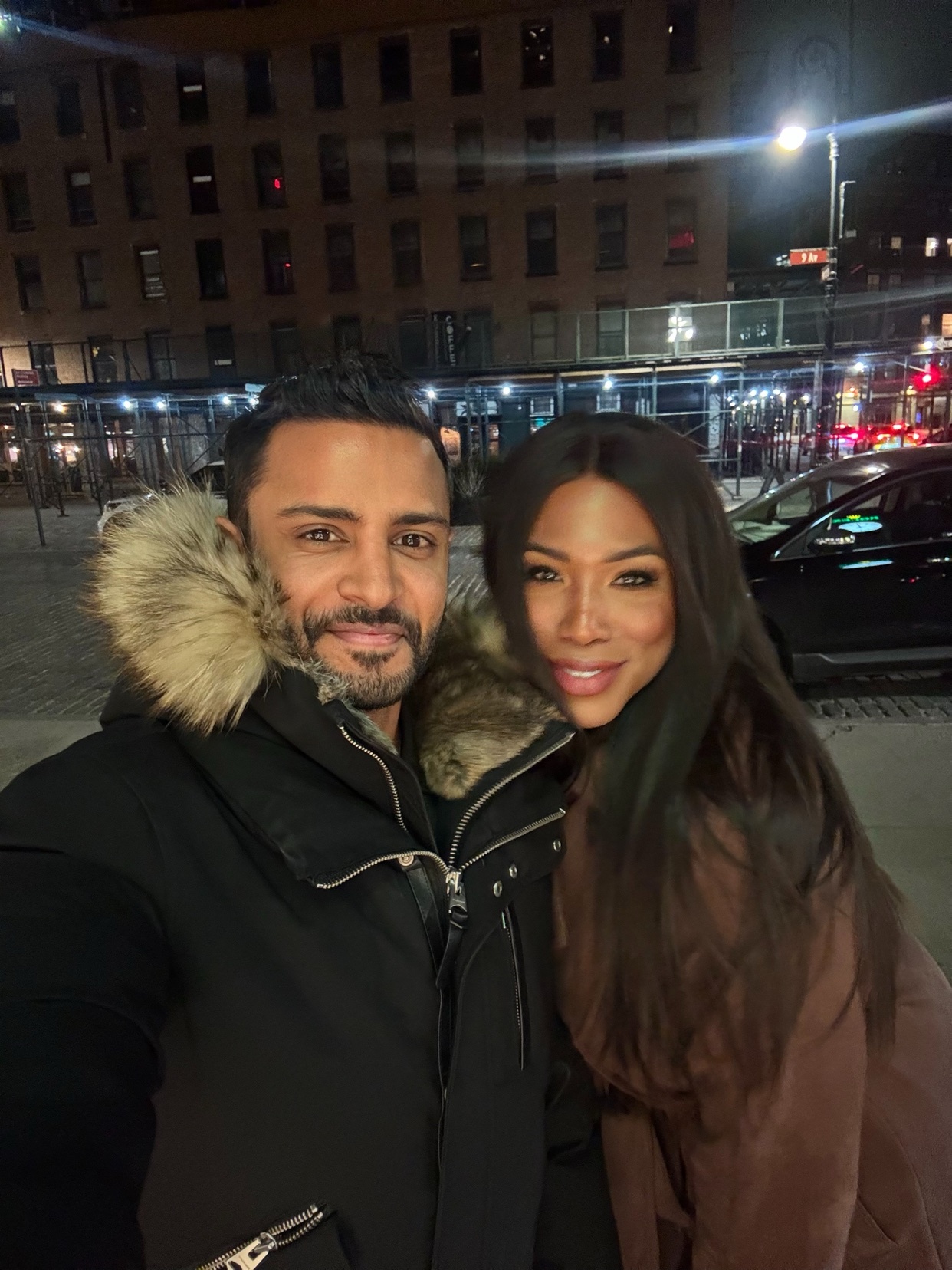 A man and woman pose for a selfie in front of a city building at night. The man wears a black winter coat with a fur-lined hood, and the woman wears a brown coat.