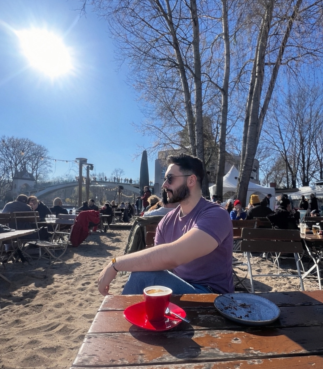 A man with a beard and sunglasses sits at a table outside a cafe with a cup of coffee in front of him. The sun is shining brightly in the clear blue sky.