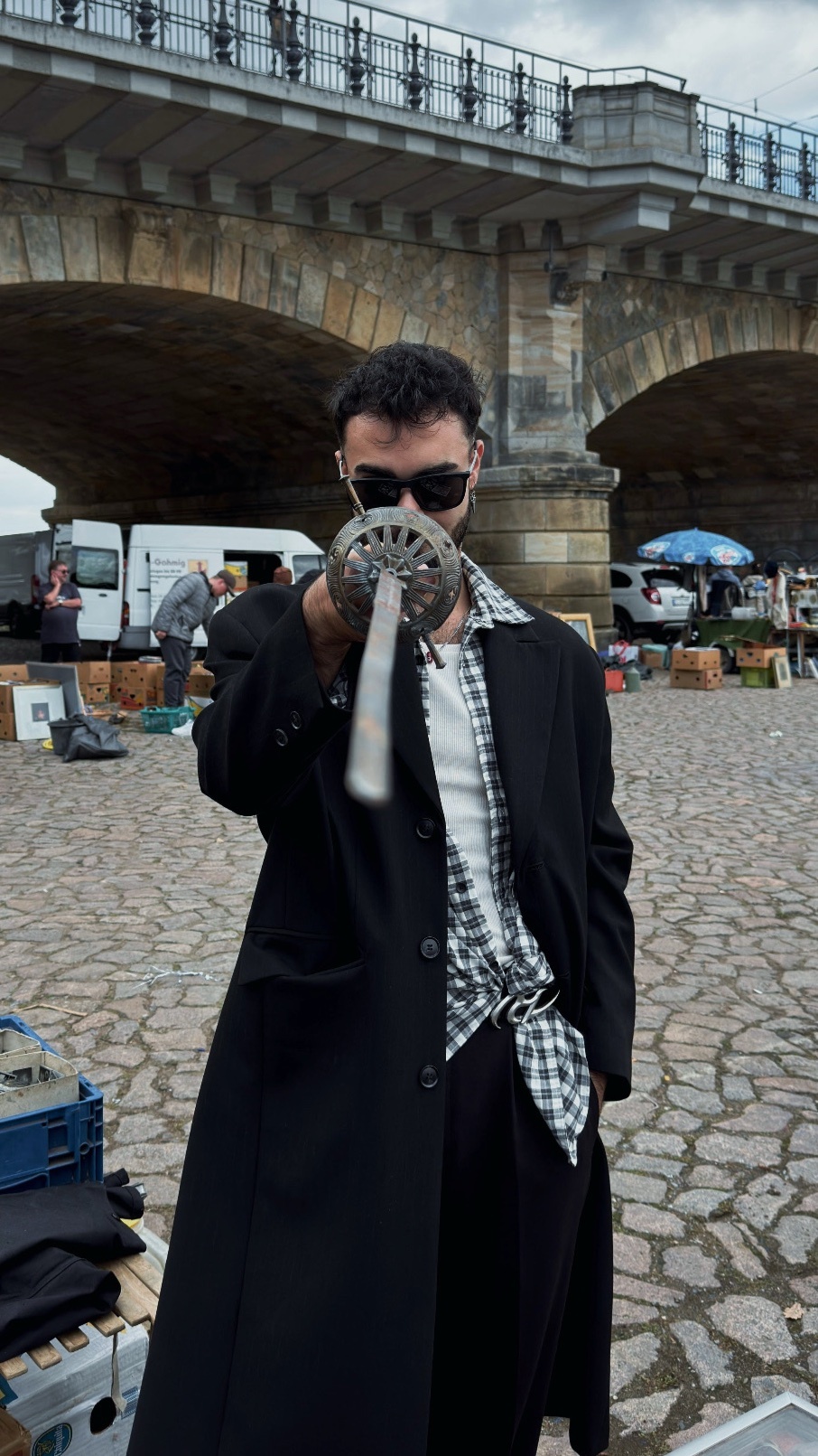 A man poses with a sword in front of a bridge. He is wearing sunglasses and a stylish black coat.